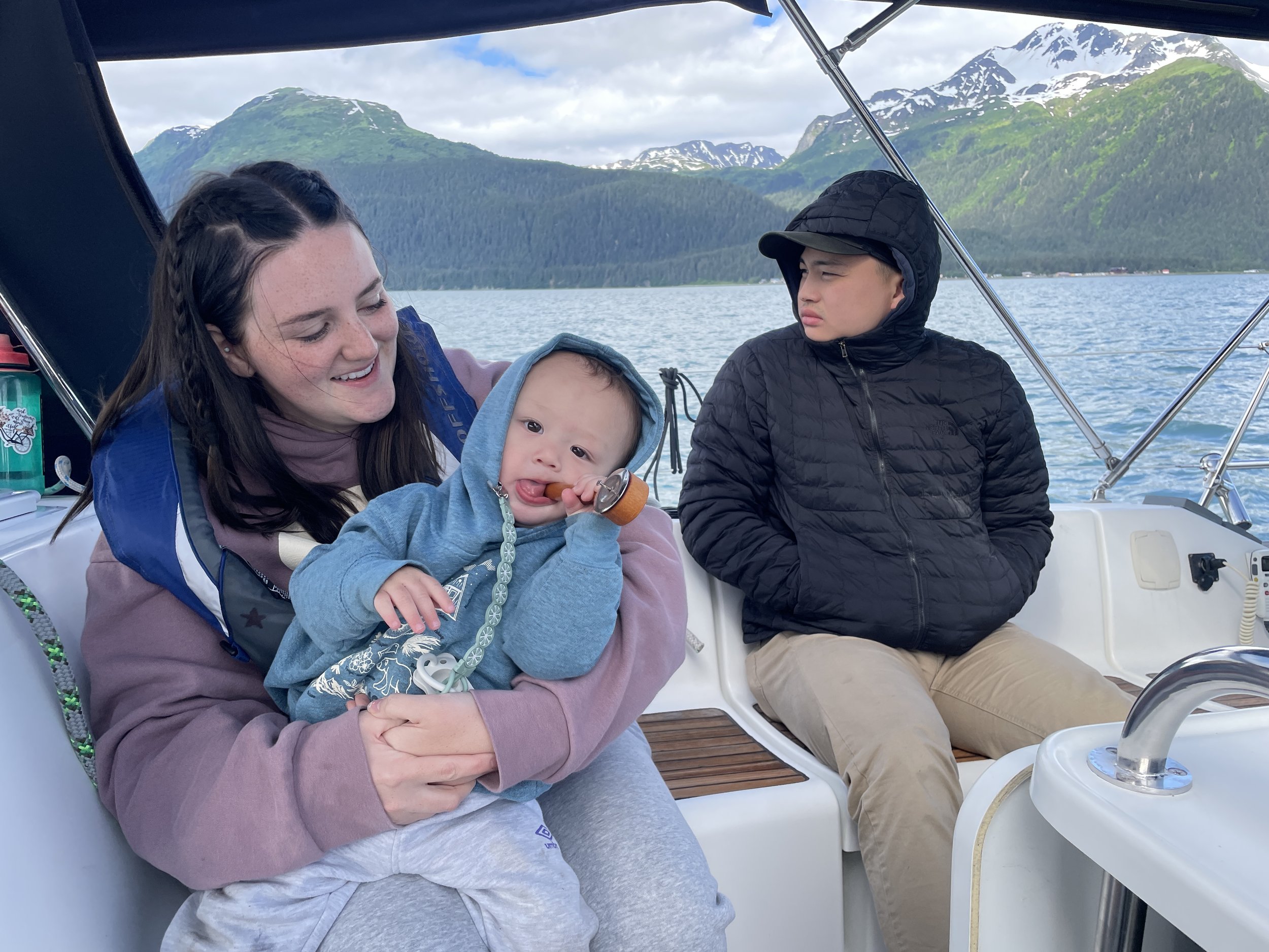 A woman holding a baby on a boat, with a young boy sitting nearby, against a backdrop of mountains and water.