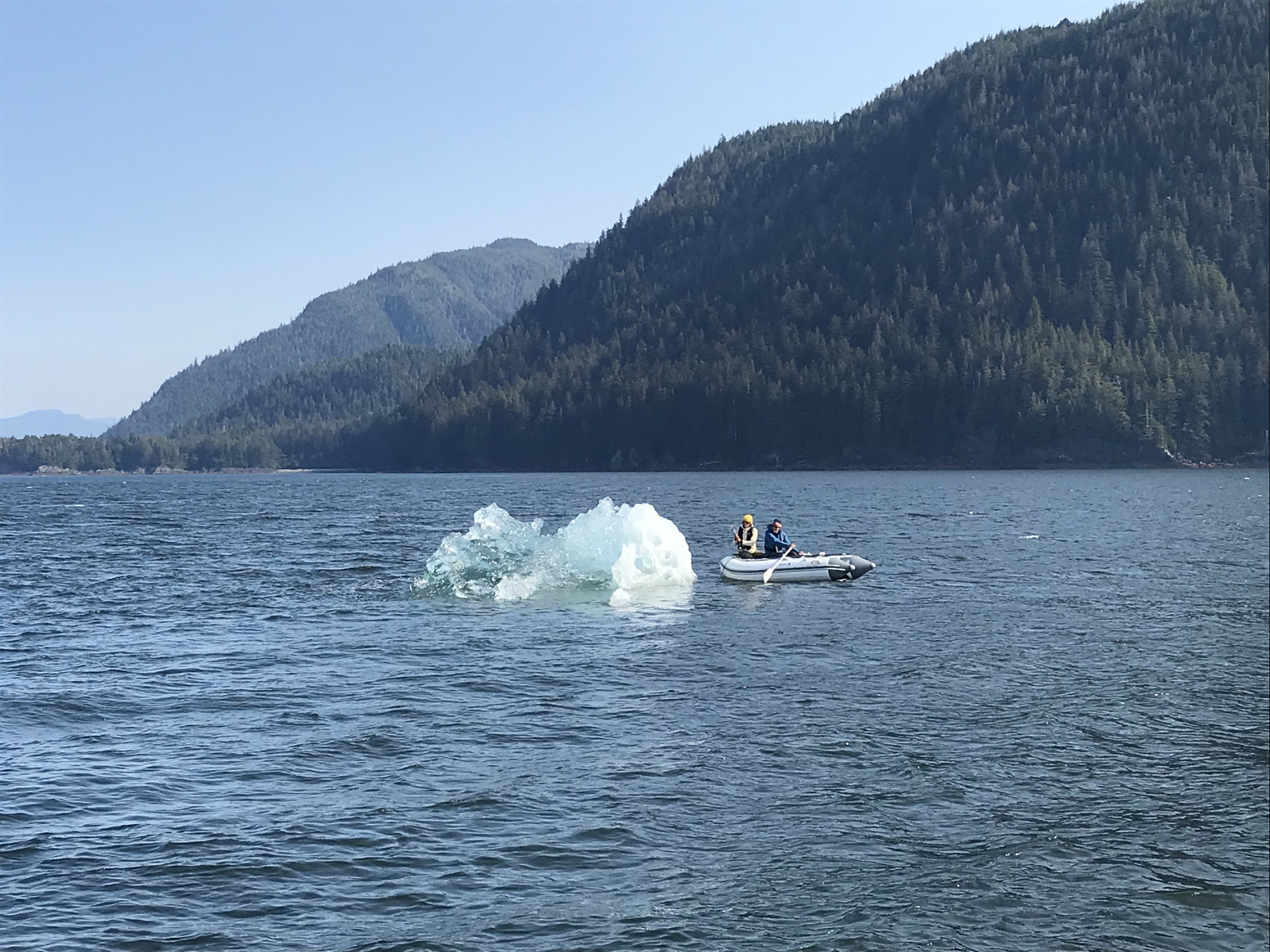 Small boat with two people near iceberg on lake surrounded by forested mountains.