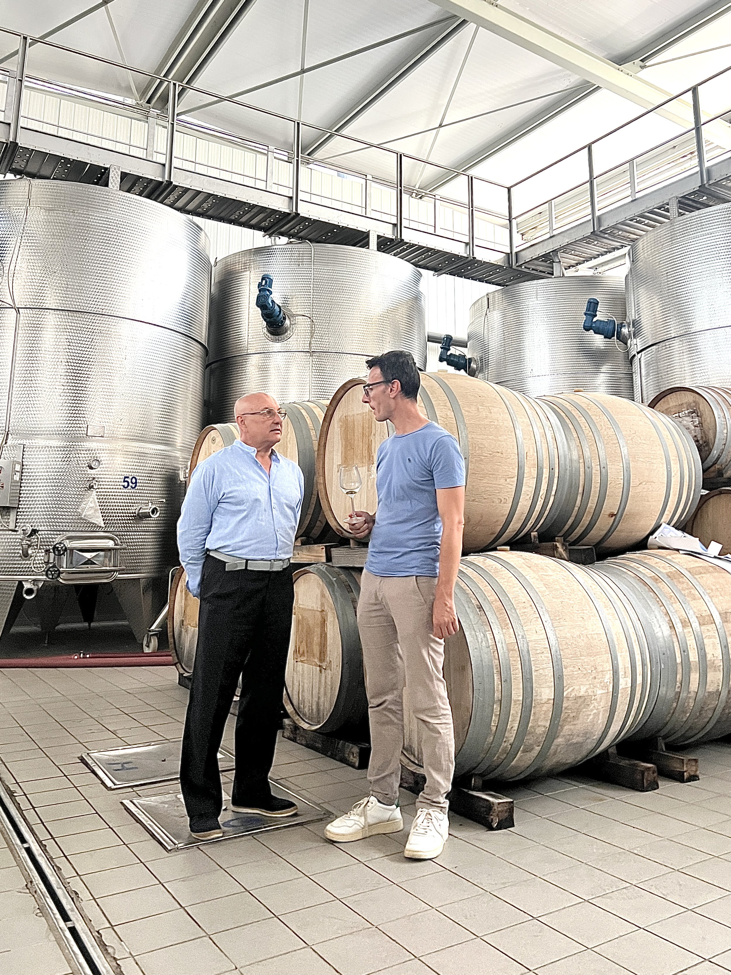 Due uomini che conversano in una cantina di vino, con grandi botti di legno e serbatoi di acciaio in background.