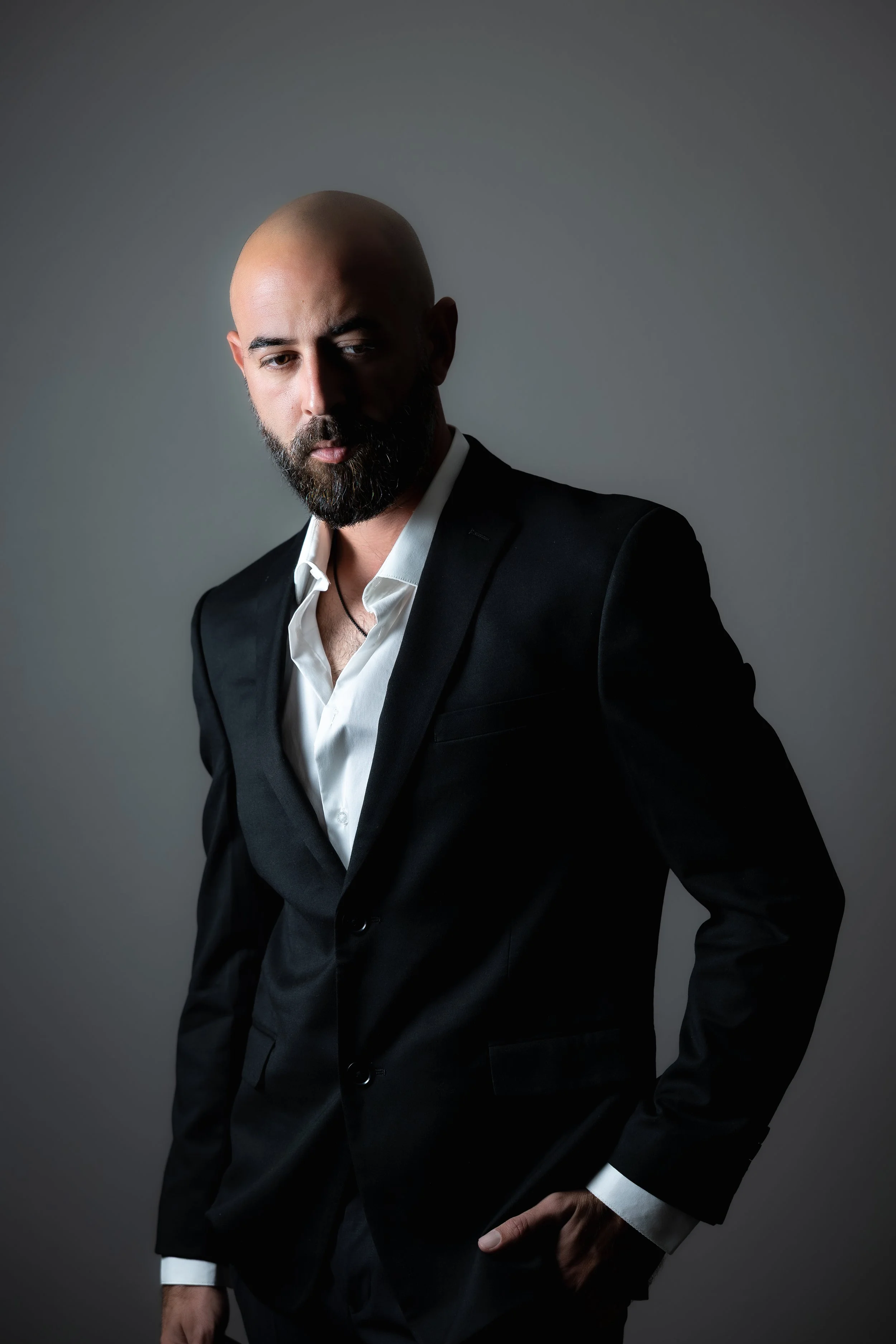 A man with a beard and bald head wearing a black suit and white shirt, standing against a plain background.