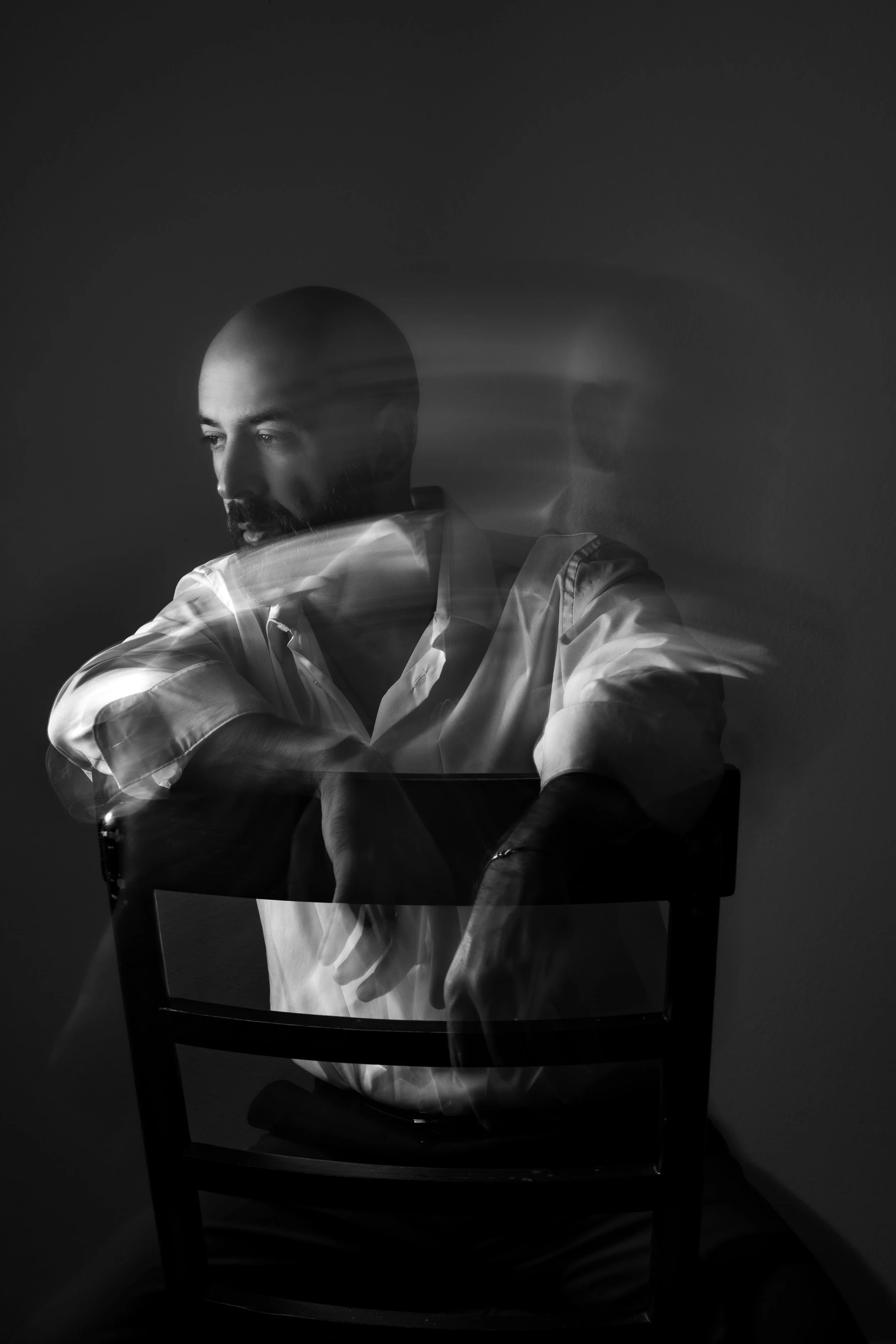 Black and white portrait of a bald man with a beard, sitting on a chair with arms crossed on a table, looking down, with motion blur and ghosting effects in a dark room.