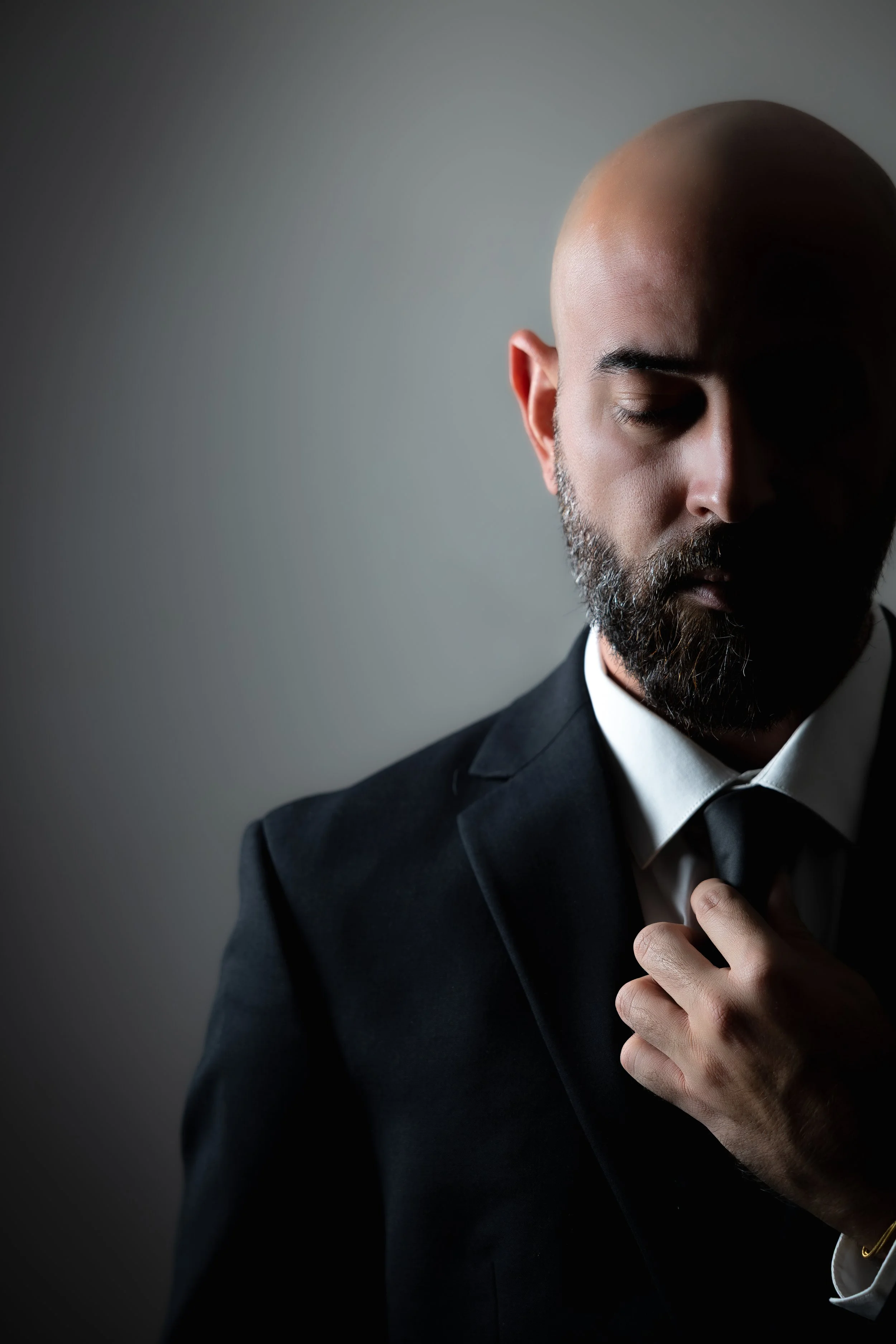 A man dressed in a black suit with a white shirt, partially adjusting his black tie, his eyes closed, with a shaved head and beard, against a plain gradient gray background.