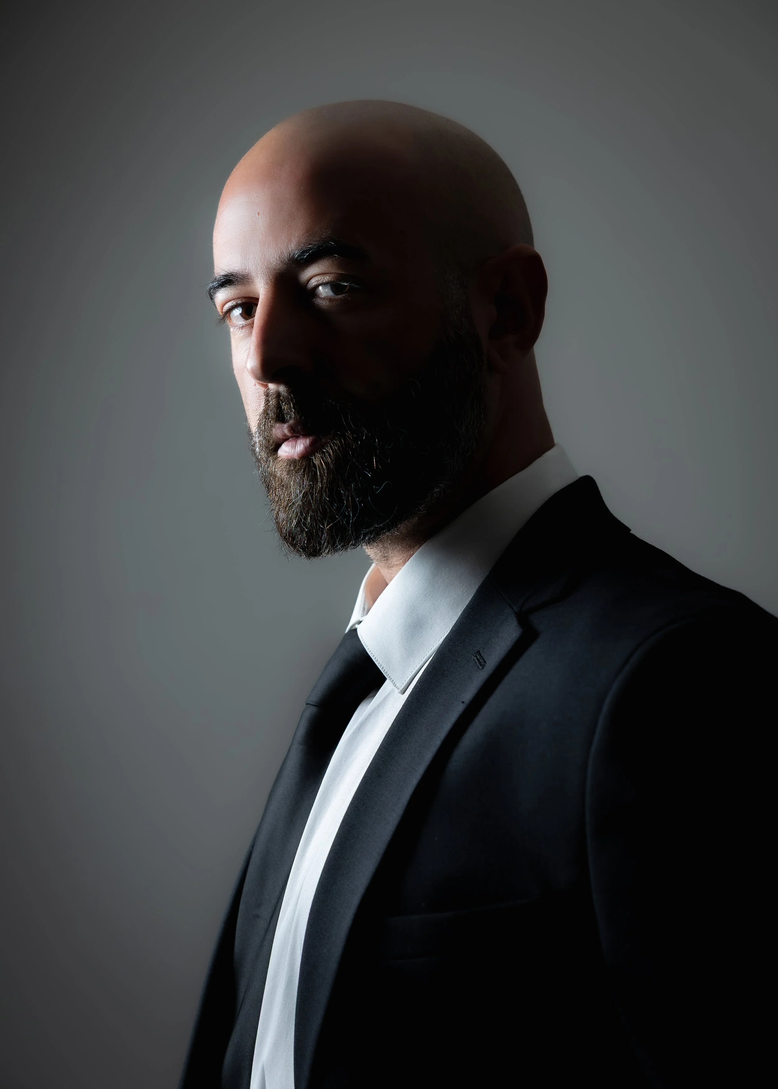 A man with a bald head and beard wearing a black tuxedo with a white shirt and black tie, looking serious, against a dark background.