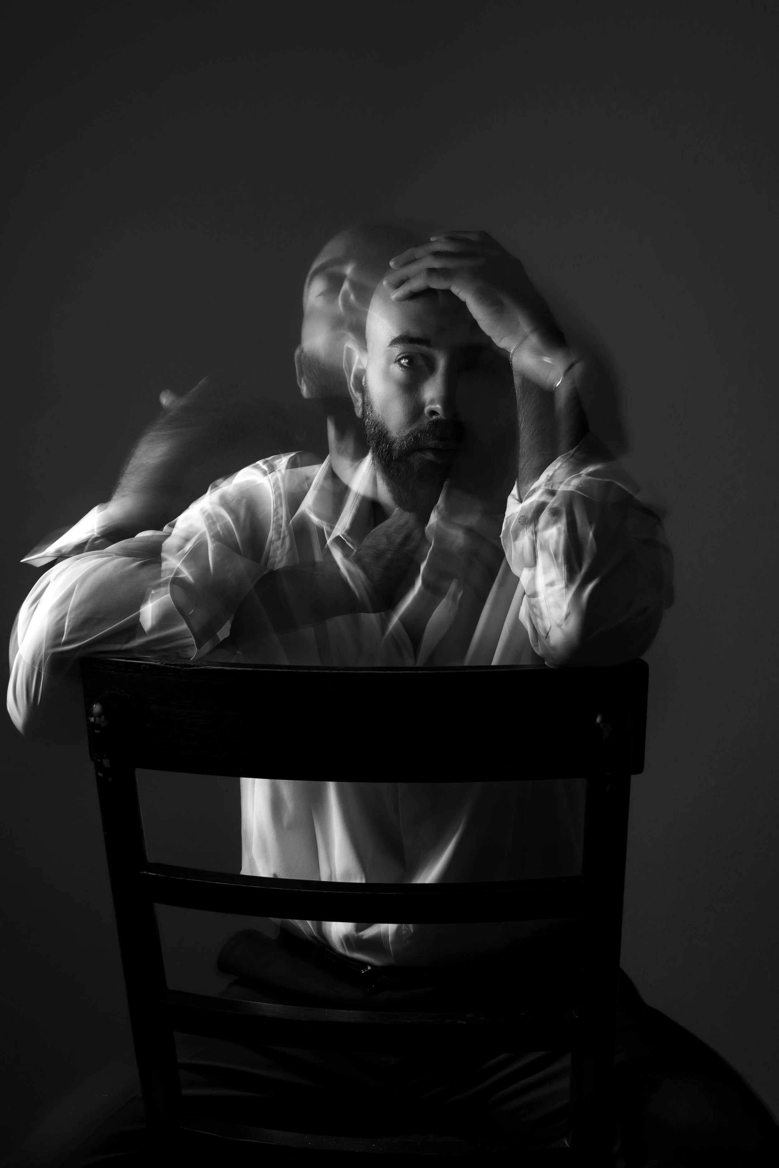 A black-and-white photo of a man sitting at a chair with multiple exposed layers showing him with different facial expressions and hand positions, creating a motion-blur effect.