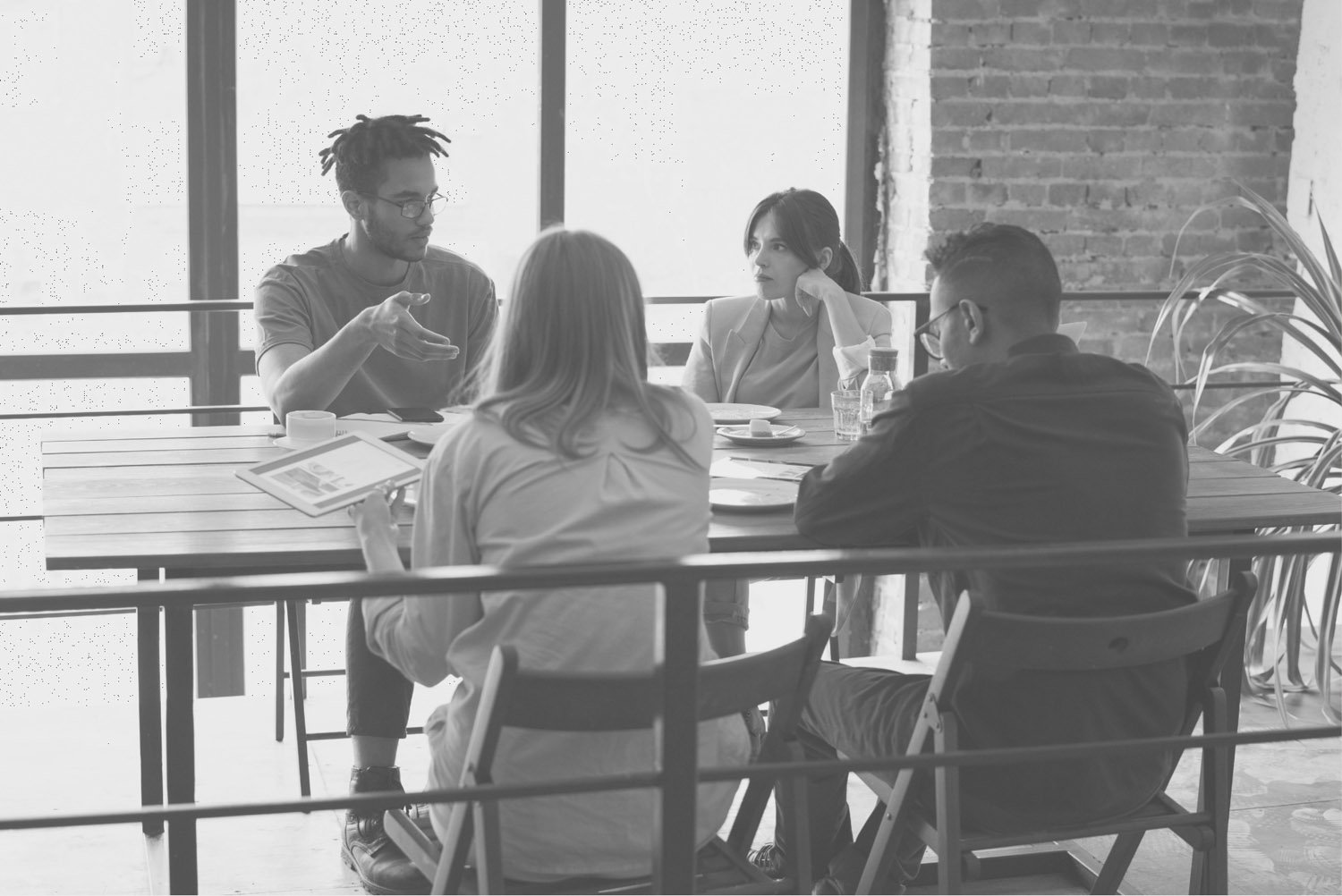 Four people sitting around a table having a discussion in a modern, industrial-style room with large windows and exposed brick walls.