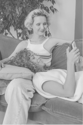 A woman sitting on a couch, smiling, while a girl with curly hair lies beside her, looking at a phone.