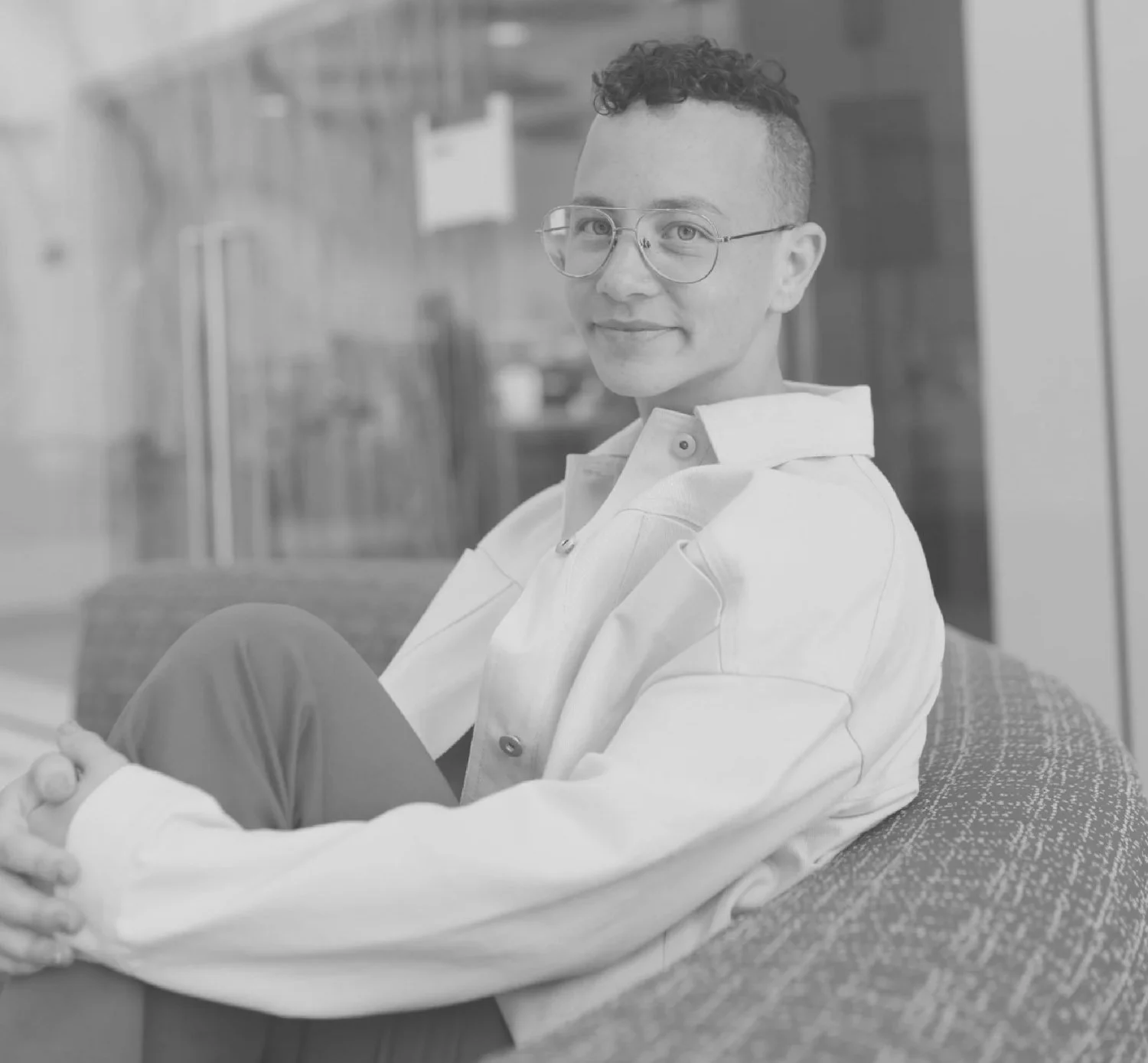 A photo of Avery with short, curly hair and glasses sitting on a couch, smiling at the camera.