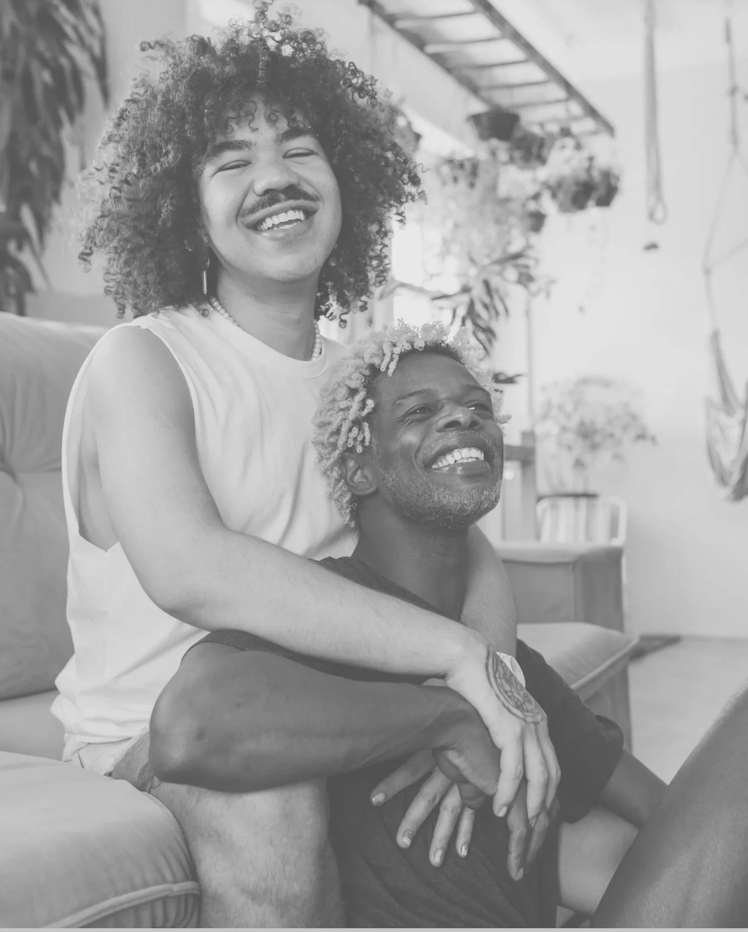 Joyful queer couple sitting together indoors, both with curly hair and smiles.