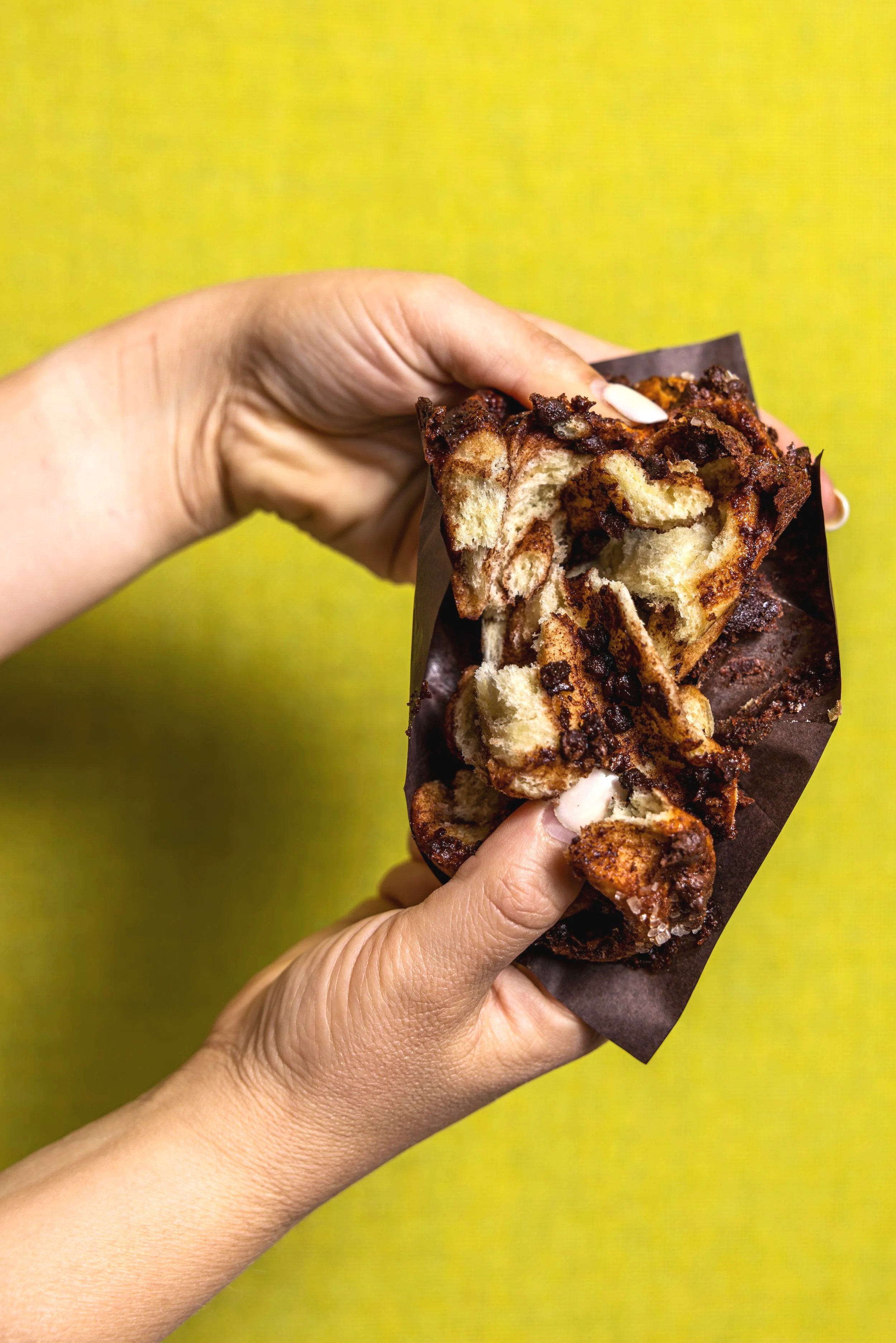 Close-up of hands holding a partially eaten chocolate chip pastry or cake with dark chocolate topping, against a lime green background.