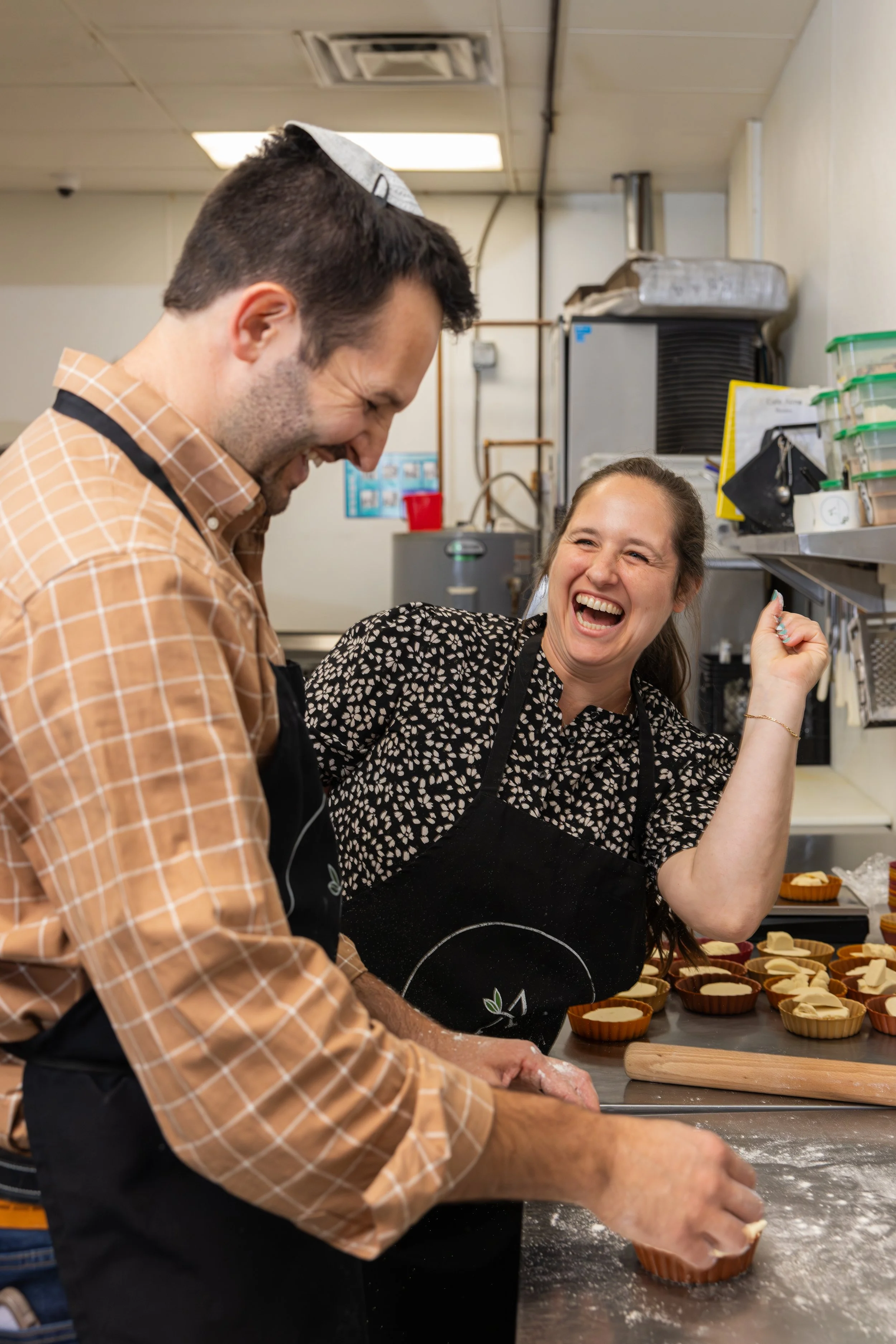 A man and woman laughing and baking cupcakes in a kitchen. The man is wearing a brown checkered shirt and a white paper hat, while the woman has a floral blouse and an apron. They are working on cupcake batter in a baking setting with cupcake liners on the counter.