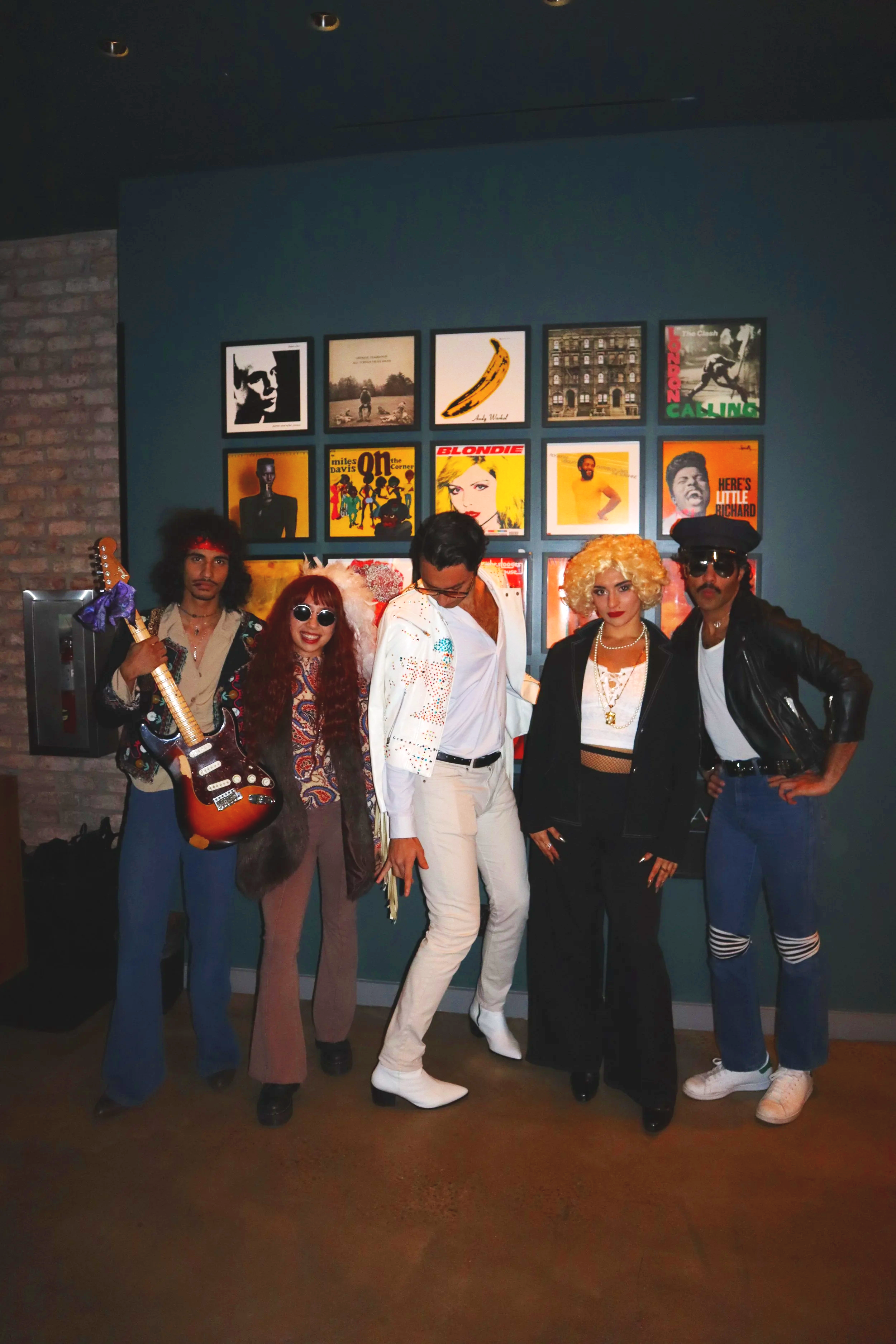 Five people dressed in colorful 1980s-style costumes standing in front of a blue wall with framed record album covers.
