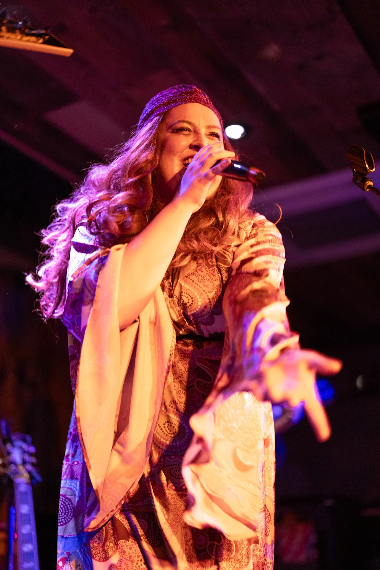 A woman with long curly hair singing into a microphone on stage, wearing a colorful patterned dress and a headband, with stage lights and musical equipment in the background.