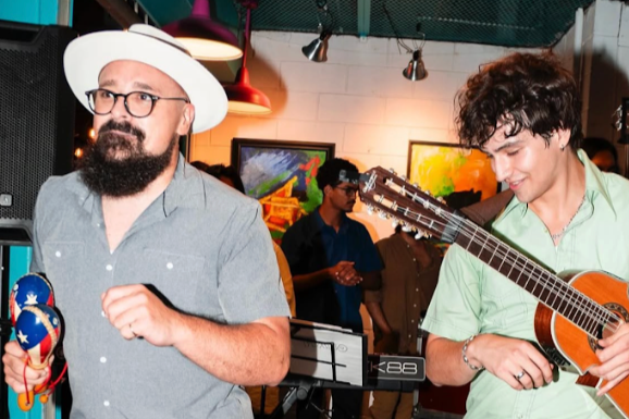 People at a social gathering, with one person playing guitar and another holding maracas, in an indoor setting with artwork on the wall.