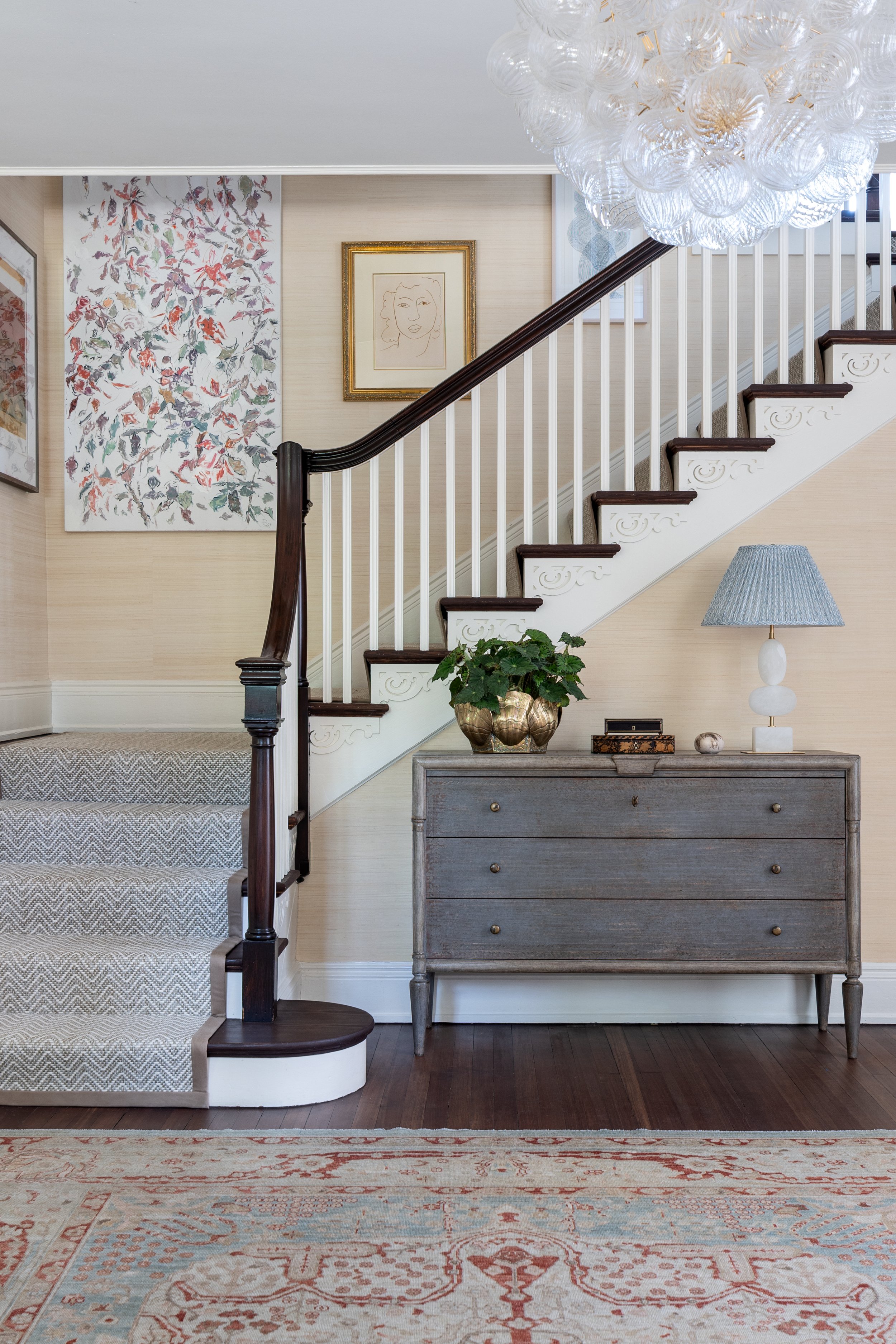 Traditional foyer with a sweeping staircase, grasscloth wallpaper, curated art gallery wall, and classic furnishings.