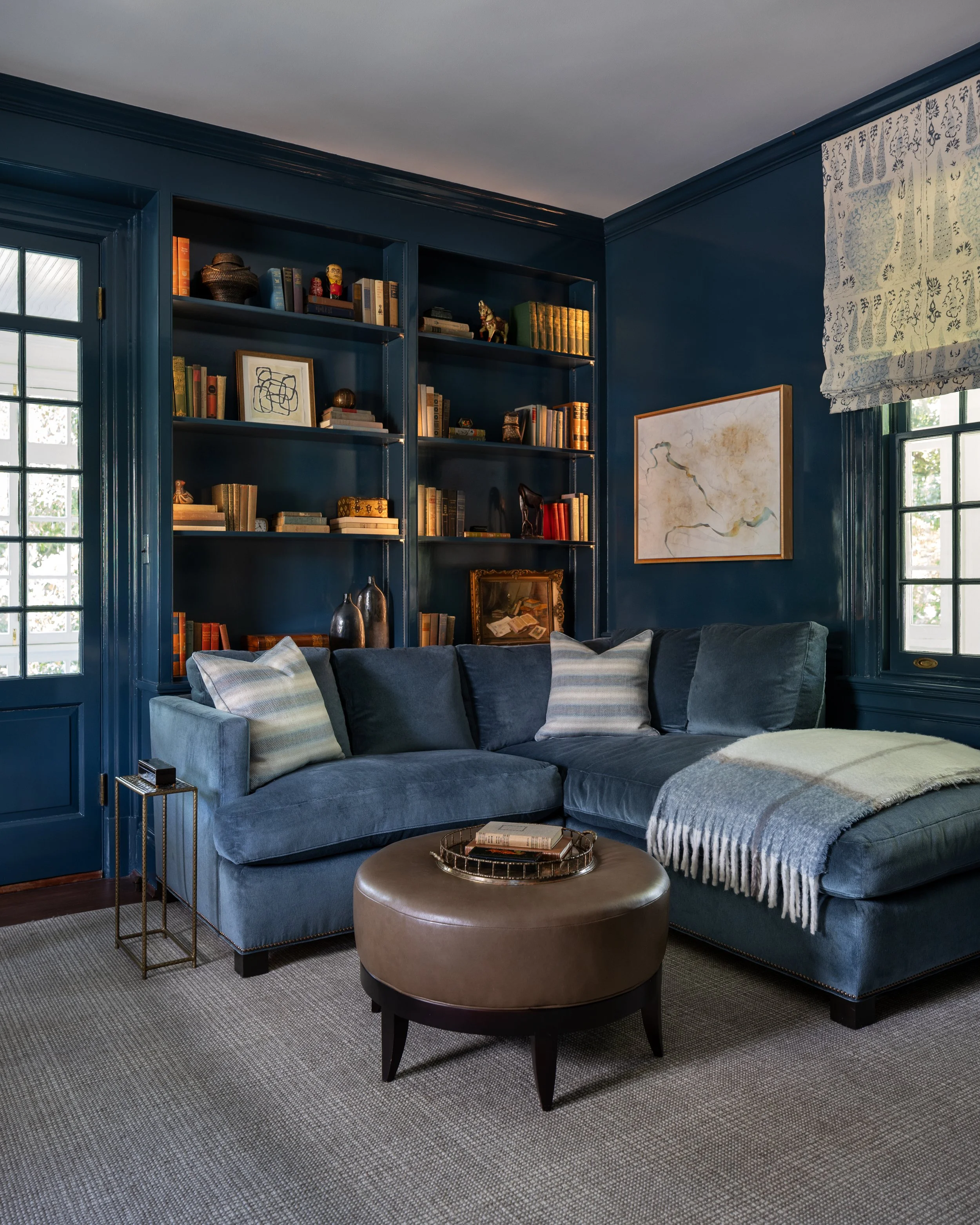 Living room with a blue sofa, patterned pillows, a round brown ottoman, a bookcase with books and decorative items, a framed artwork, and windows with patterned Roman shades.