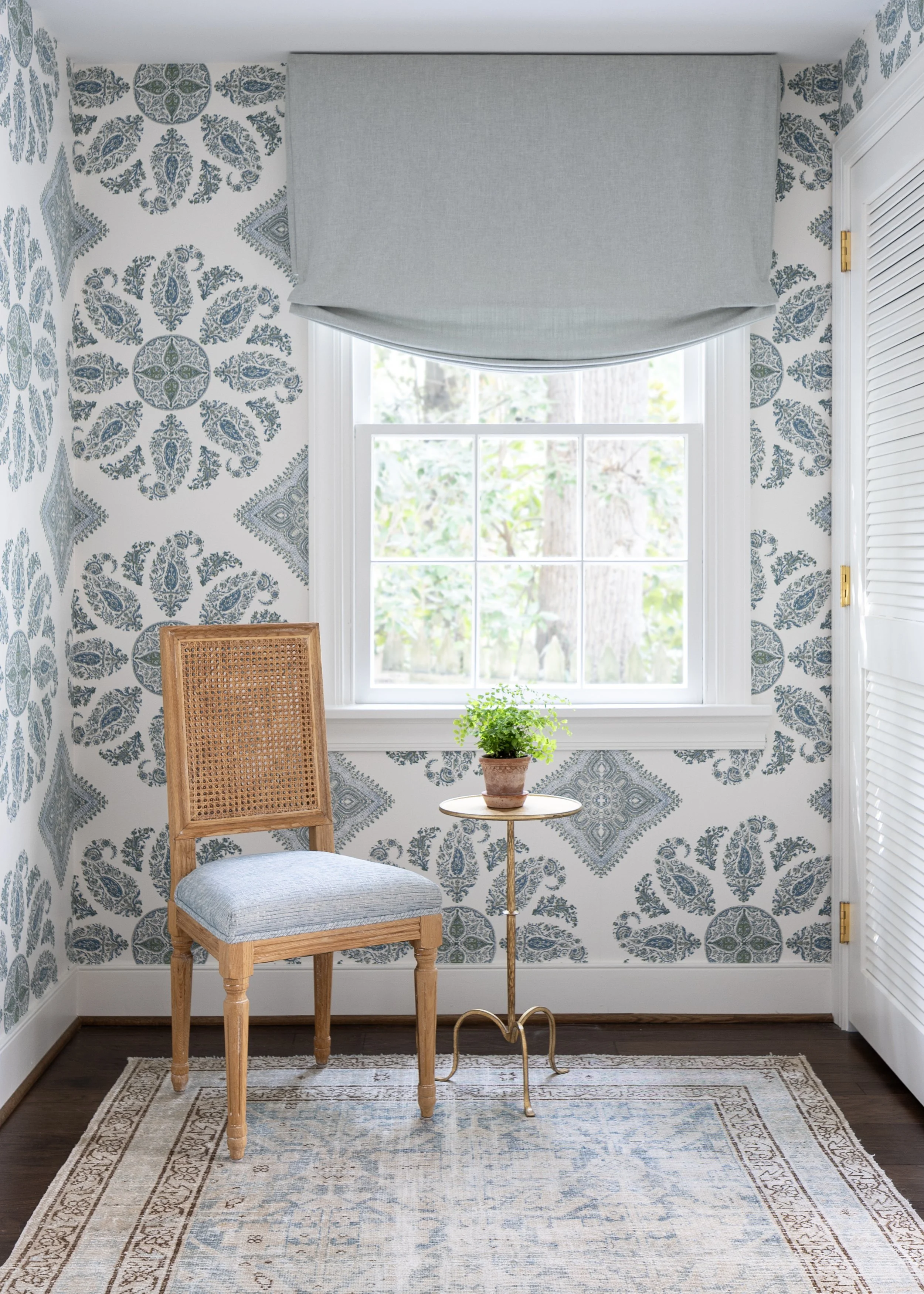 A corner of a room with blue and white patterned wallpaper, a window with a light gray valance, a wooden chair with a woven back and a cushioned seat, a small round table with a potted green plant, and a beige patterned rug on dark hardwood floor.