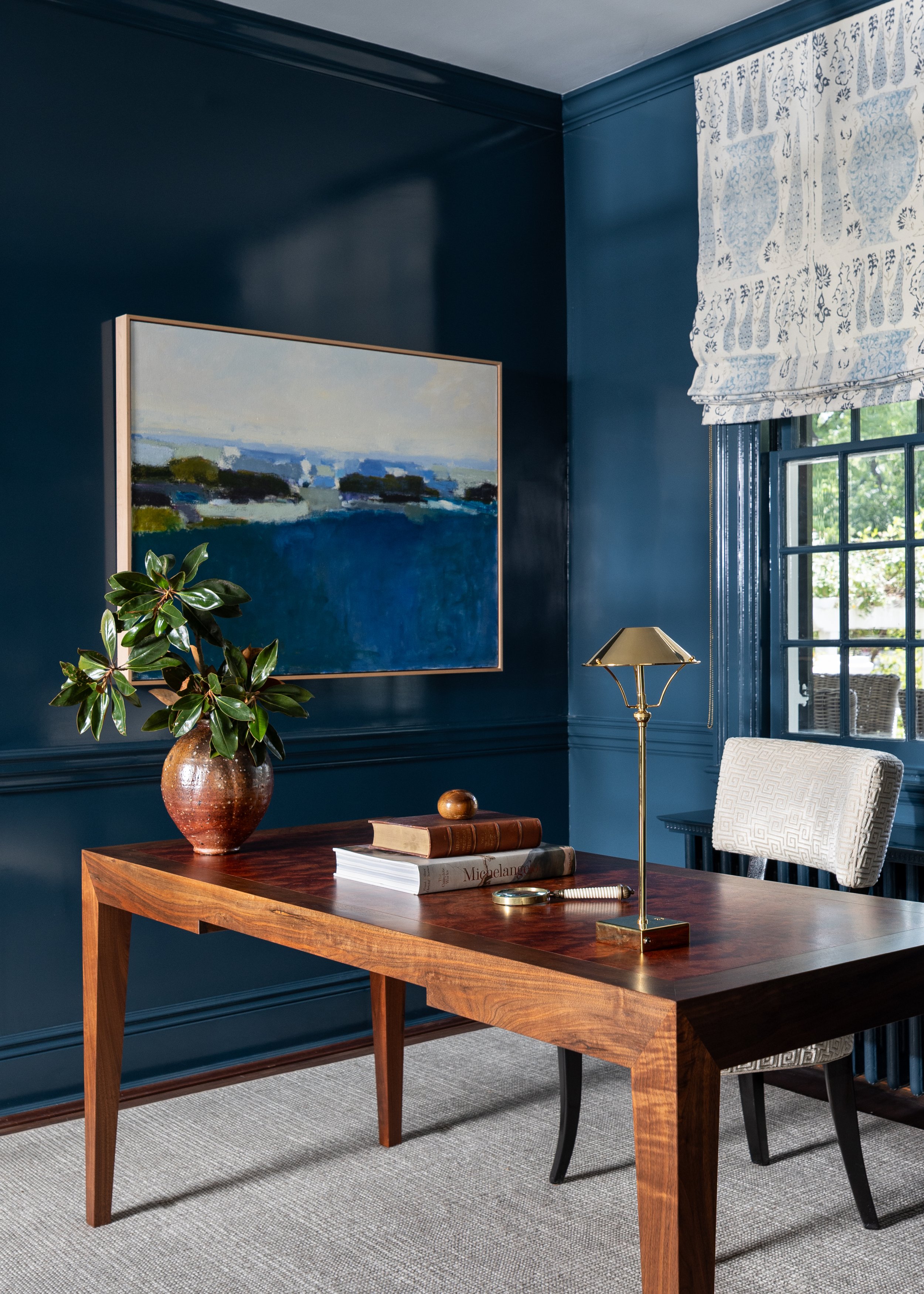 Home office vignette with deep blue walls, custom desk, original artwork, bookcases, and natural light.