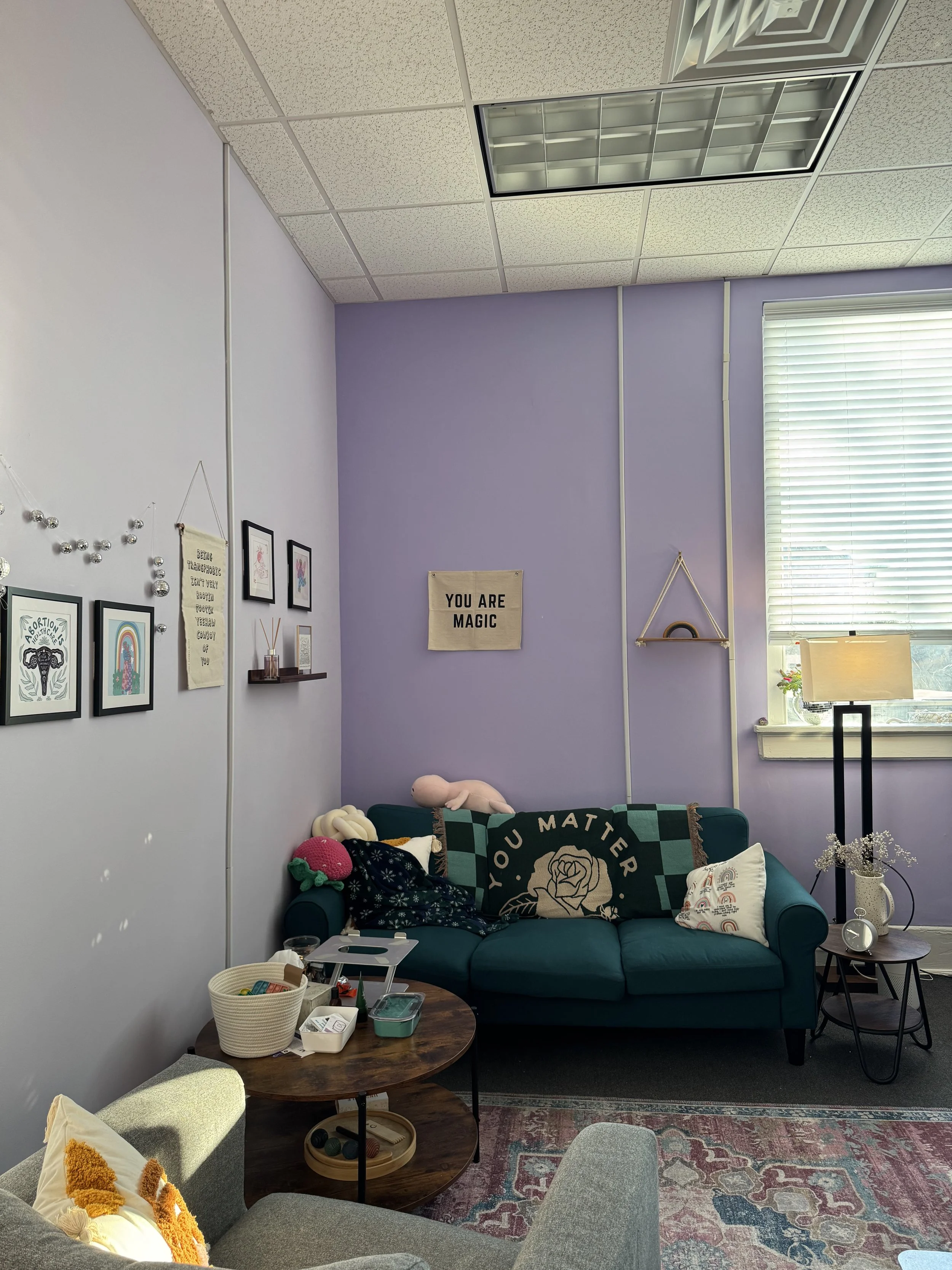 A cozy room corner with purple walls, a teal sofa with stuffed animals and cushions, a wooden side table with decorations, and a window with blinds letting in natural light. Decor includes framed art, a sign reading 'YOU ARE MAGIC,' and a floor lamp.