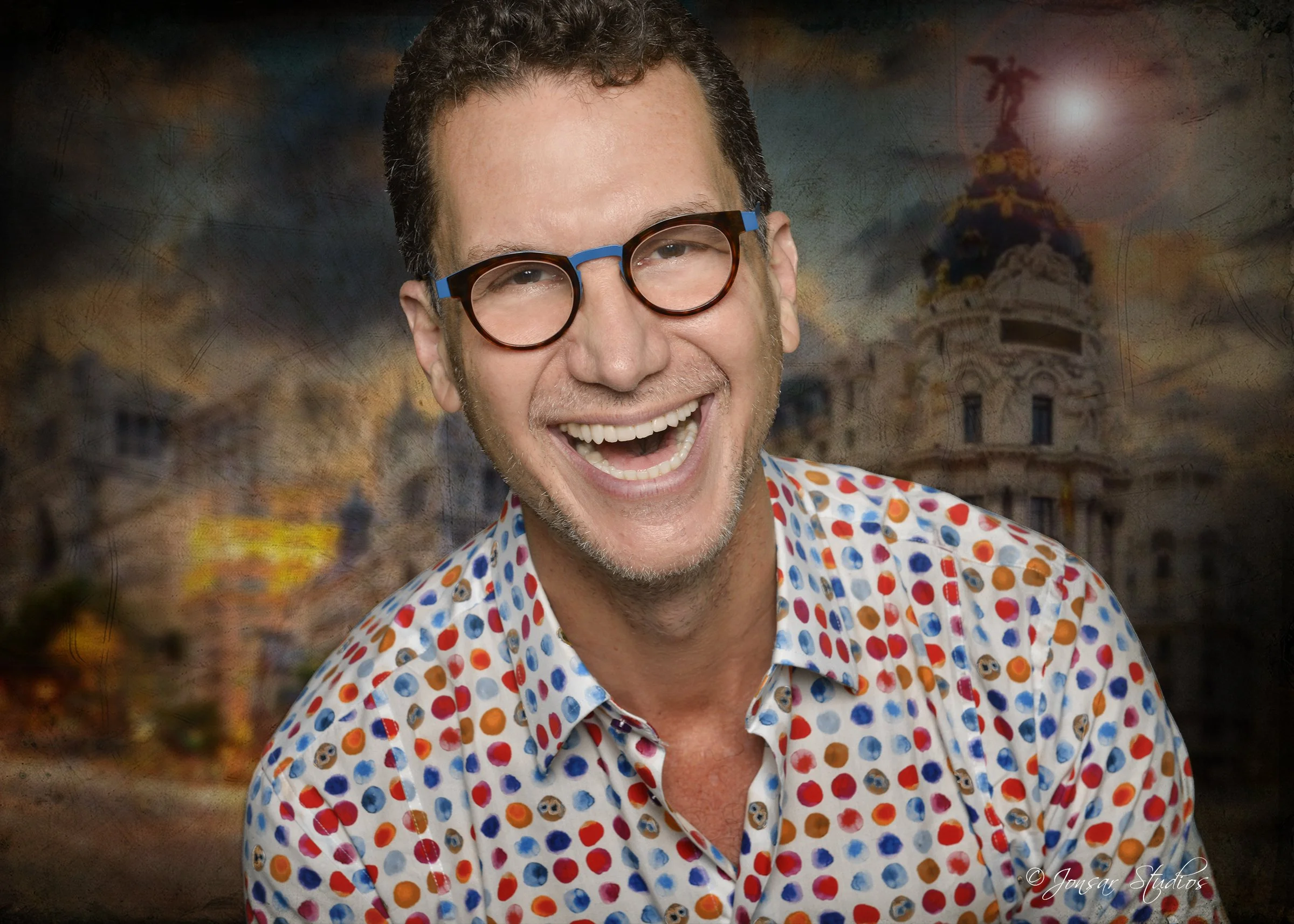 A smiling Steven Shulem wearing glasses and a colorful polka dot shirt in front of a blurred, historical building.