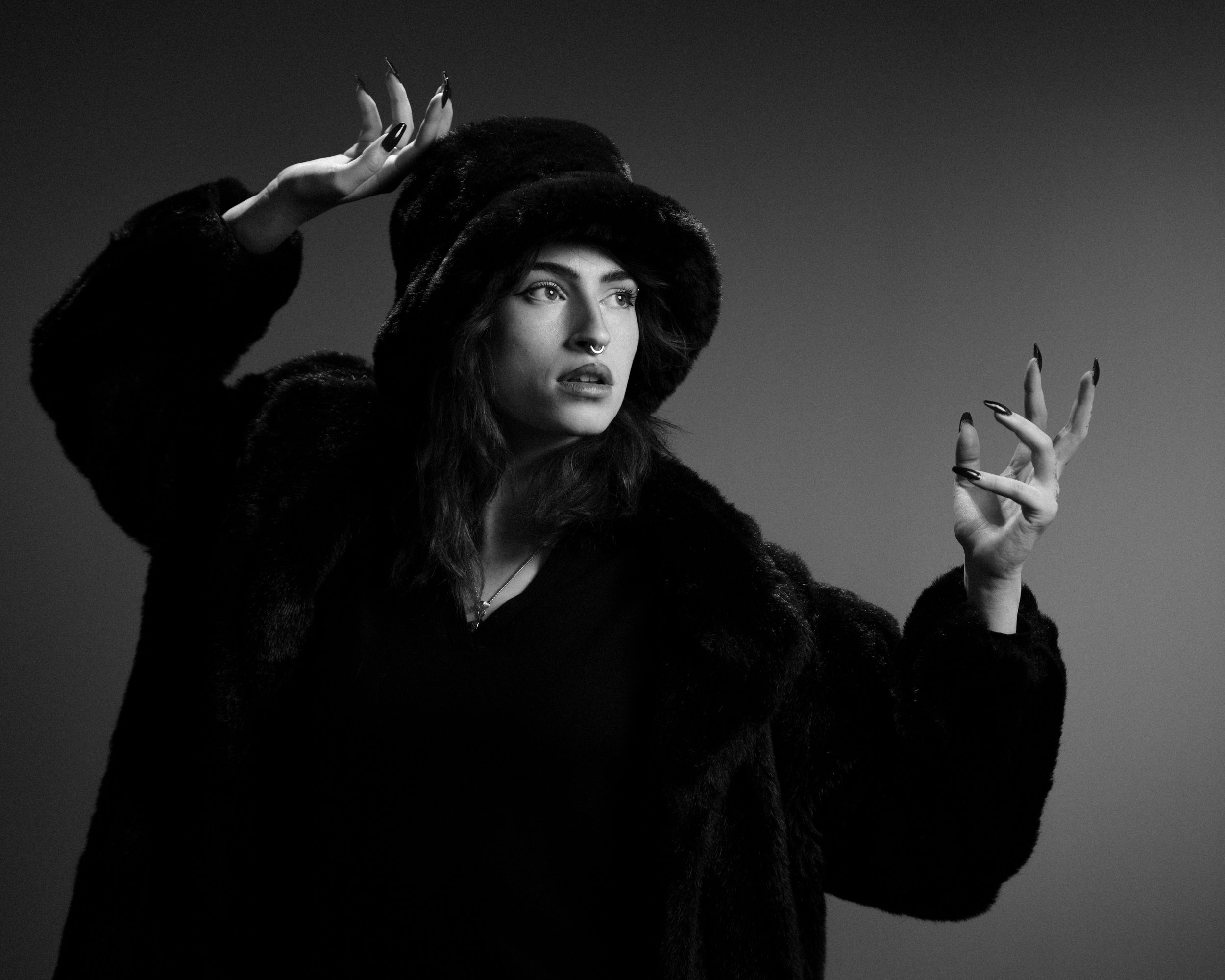 A woman wearing a fur coat and hat poses with her hands raised against a plain background in black and white.
