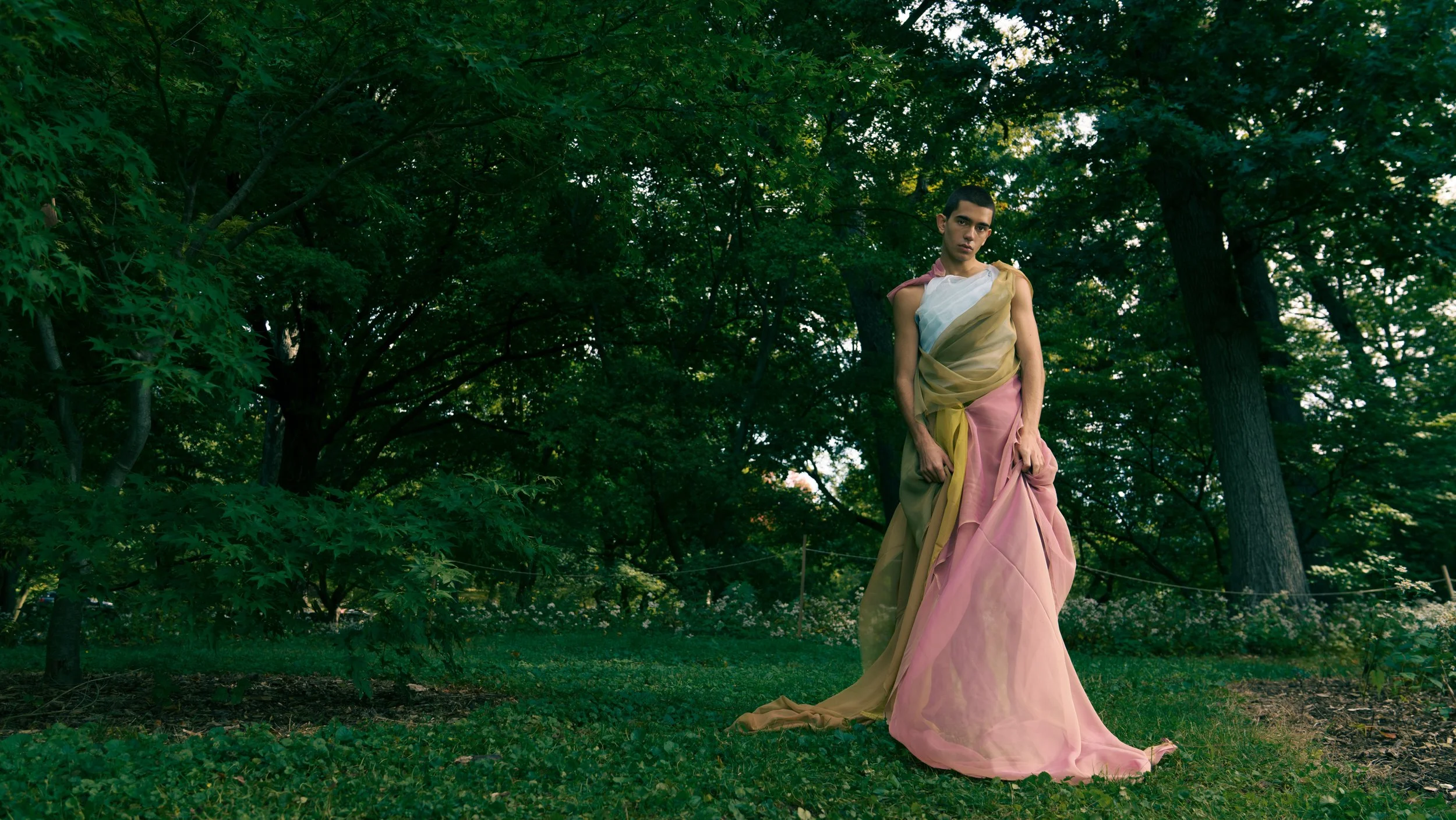 A person standing outdoors in a lush green wooded area, wearing a long, flowing, multicolored dress with shades of pink, yellow, and white.