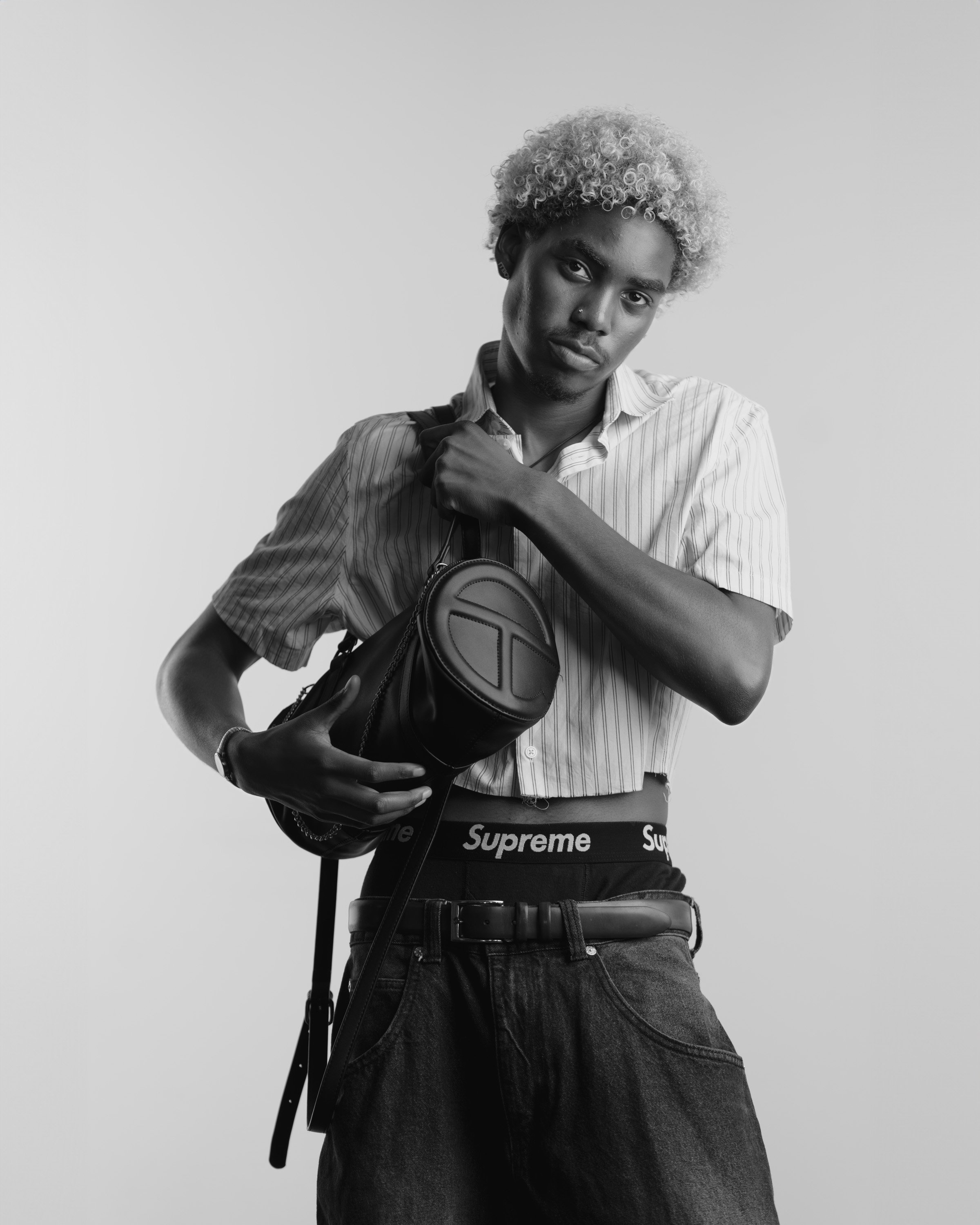 A young person with curly hair wearing a striped shirt and Supreme brand underwear, holding a small bag, posing against a plain background.