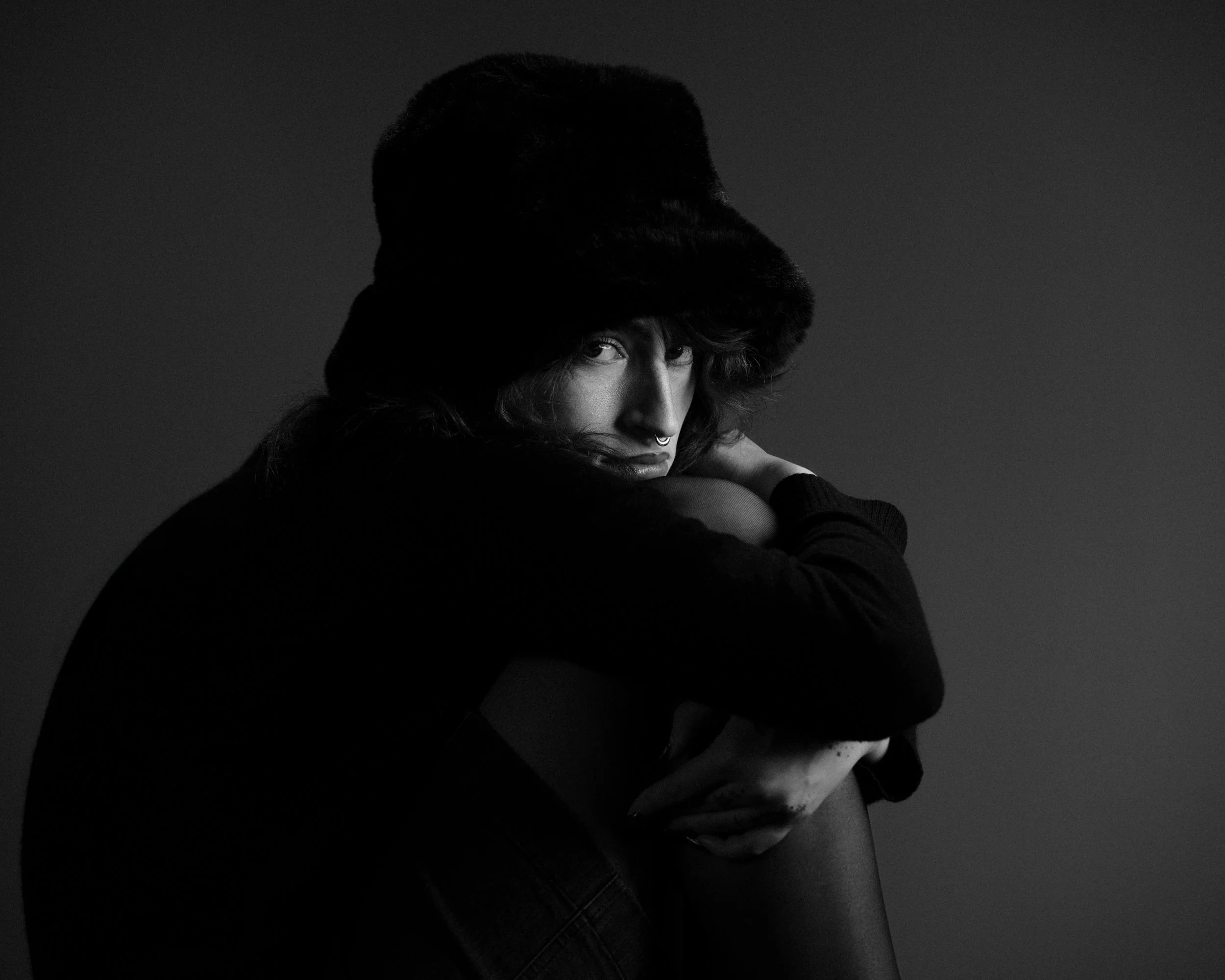 Black and white photo of a person sitting with their knees drawn up, wearing a large fur hat and dark clothing, gazing intently at the camera.