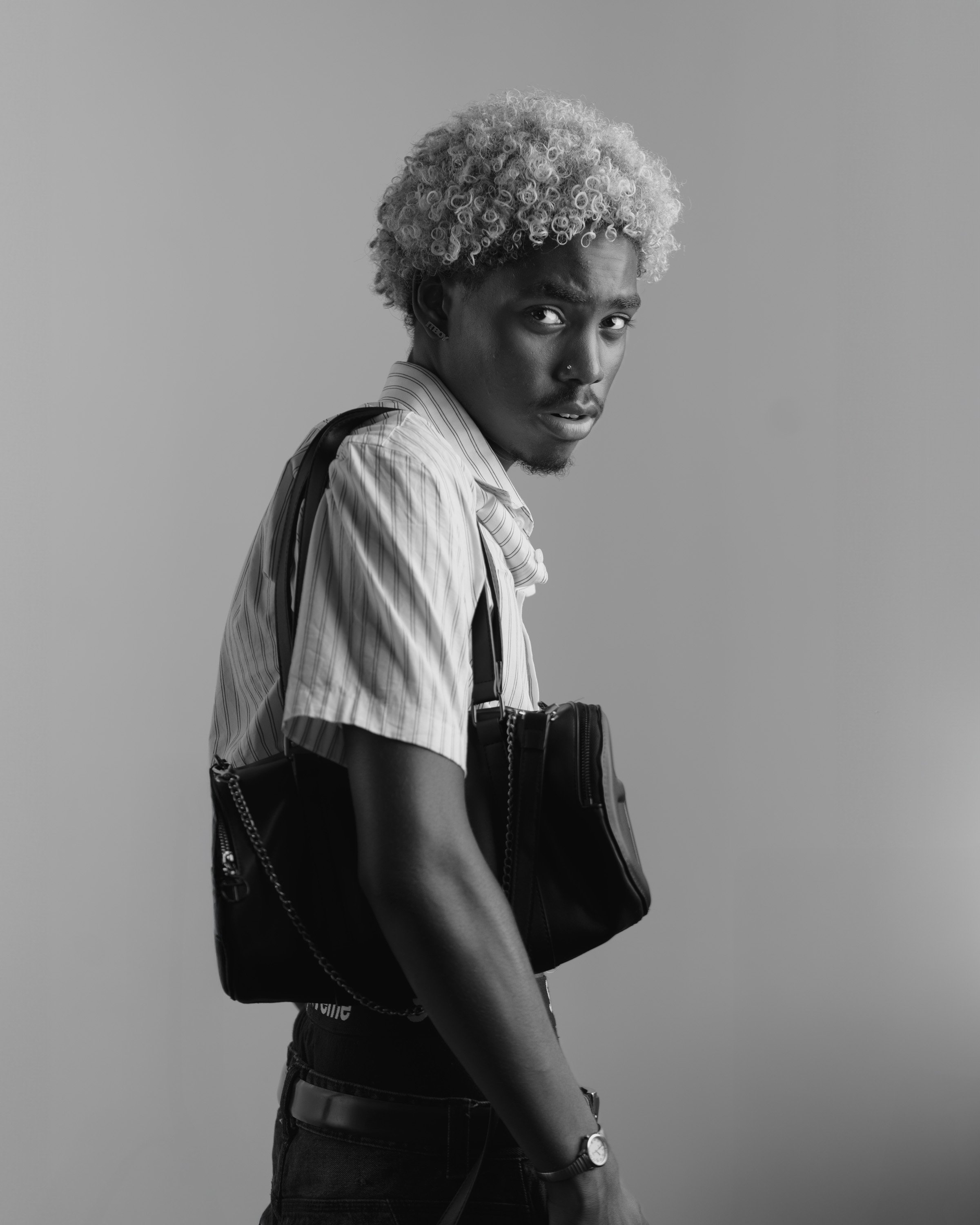 Black and white photo of a young person with curly hair, wearing a striped shirt, carrying a shoulder bag, and looking over their shoulder.