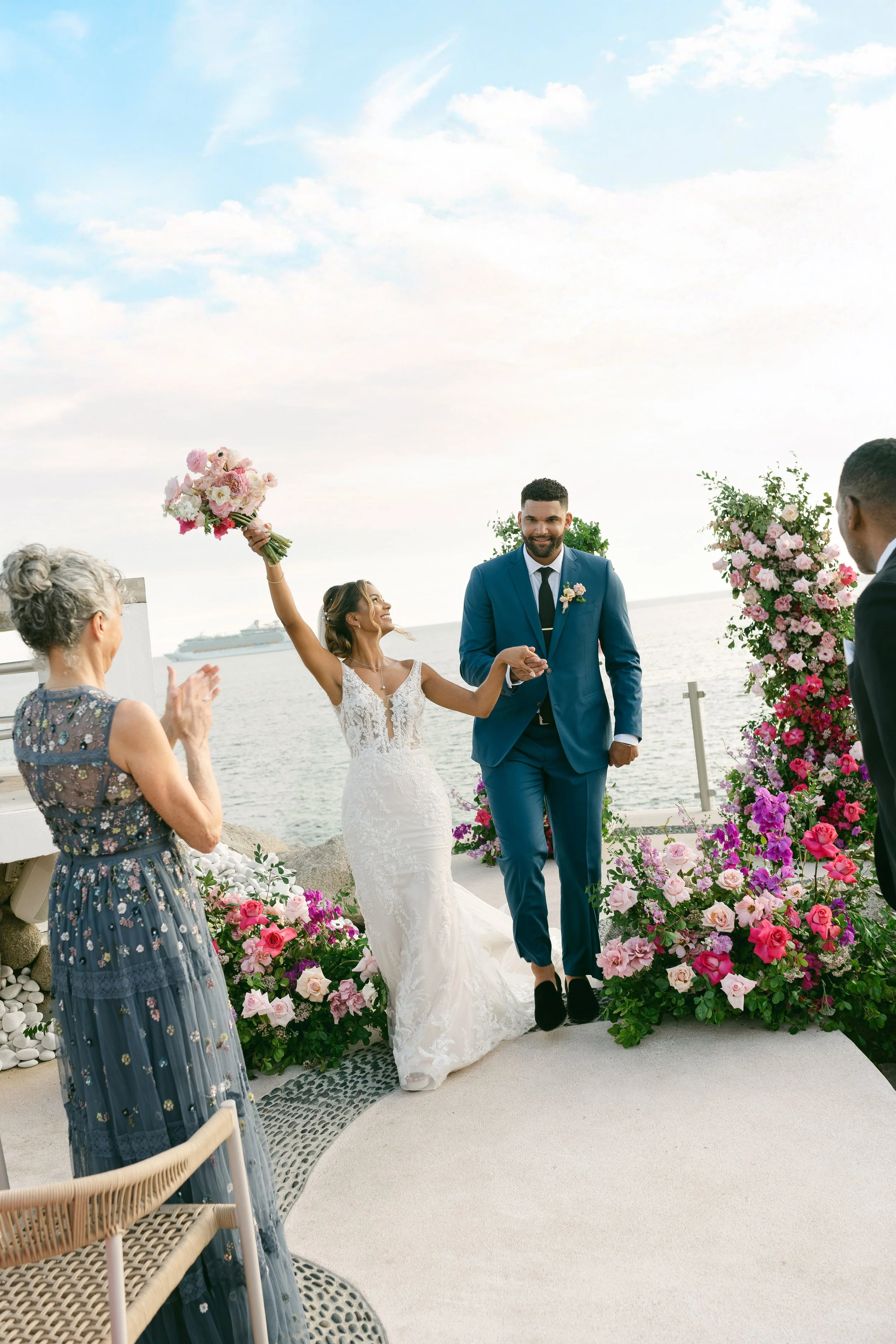 Pareja de novios caminando juntos en una ceremonia al aire libre junto al mar, rodeados de flores en tonos rosados y blancos, con invitados aplaudiendo y un crucero en el agua al fondo.