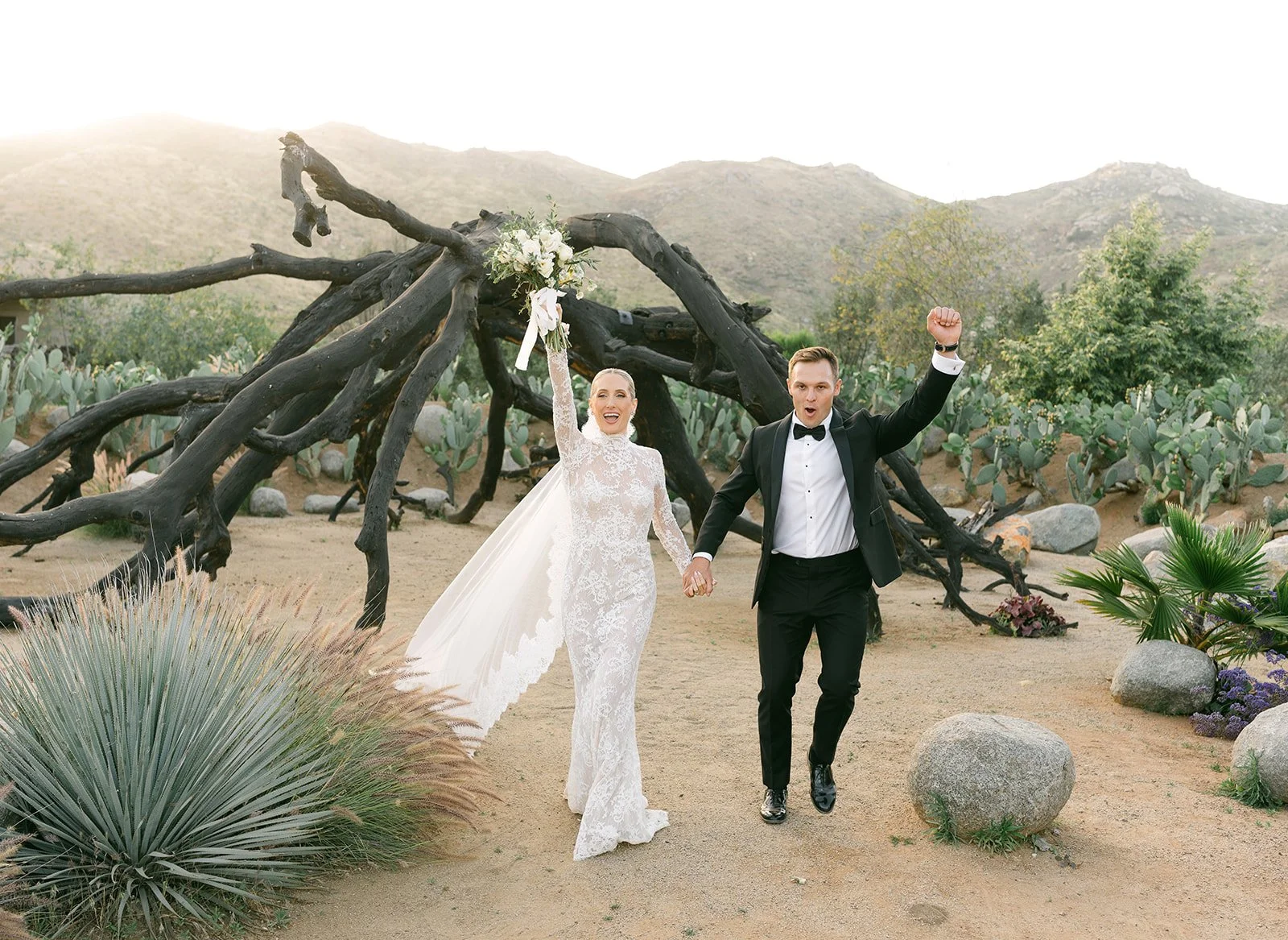 Pareja de recién casados caminando en un paisaje desértico con cactus y plantas. La mujer lleva vestido de novia y sostiene ramo de flores, el hombre viste traje formal. Ambos parecen felices y celebran su boda.
