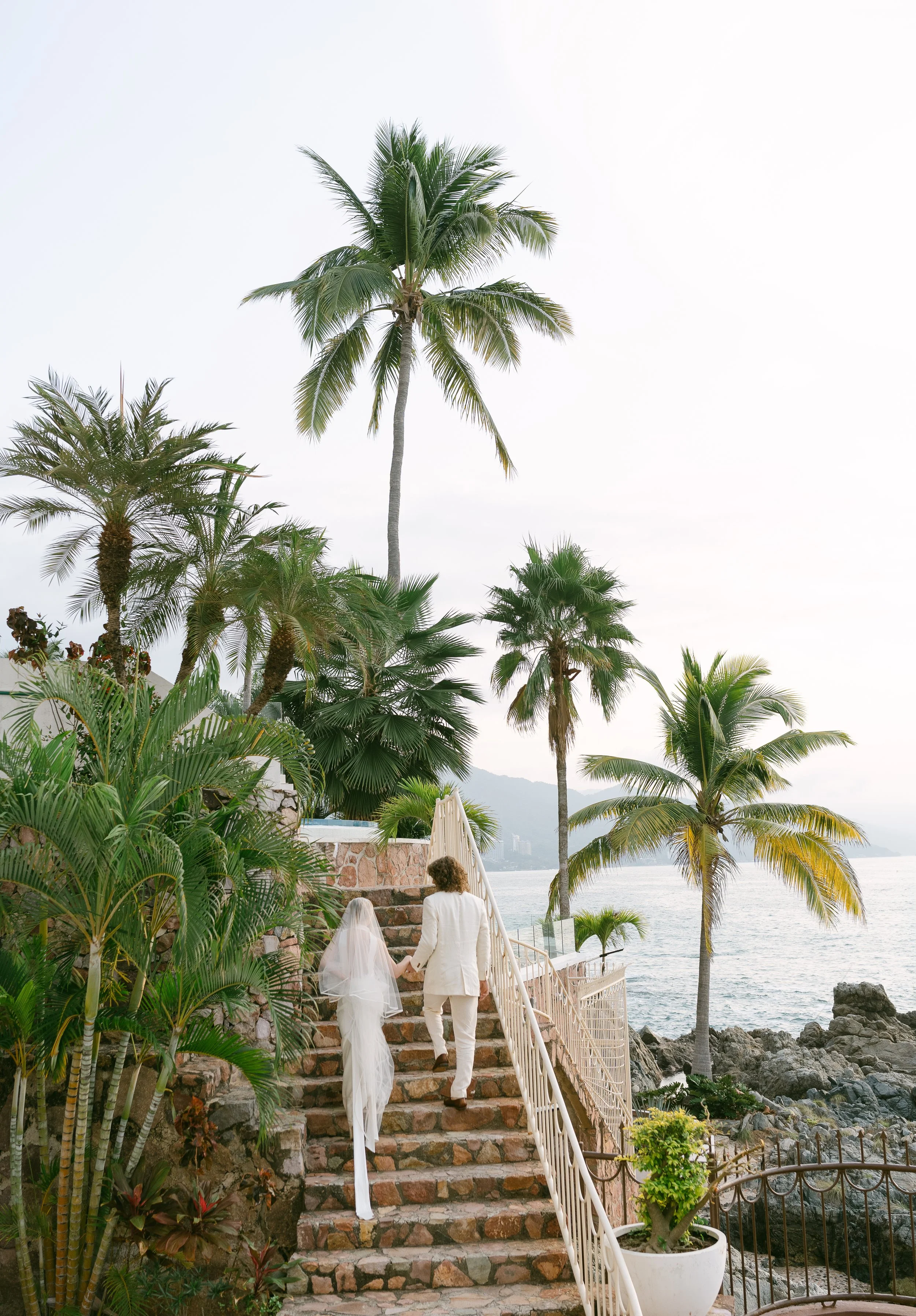 Pareja caminando por una escalera junto a la playa, rodeados de palmeras y vegetación tropical en un entorno costero