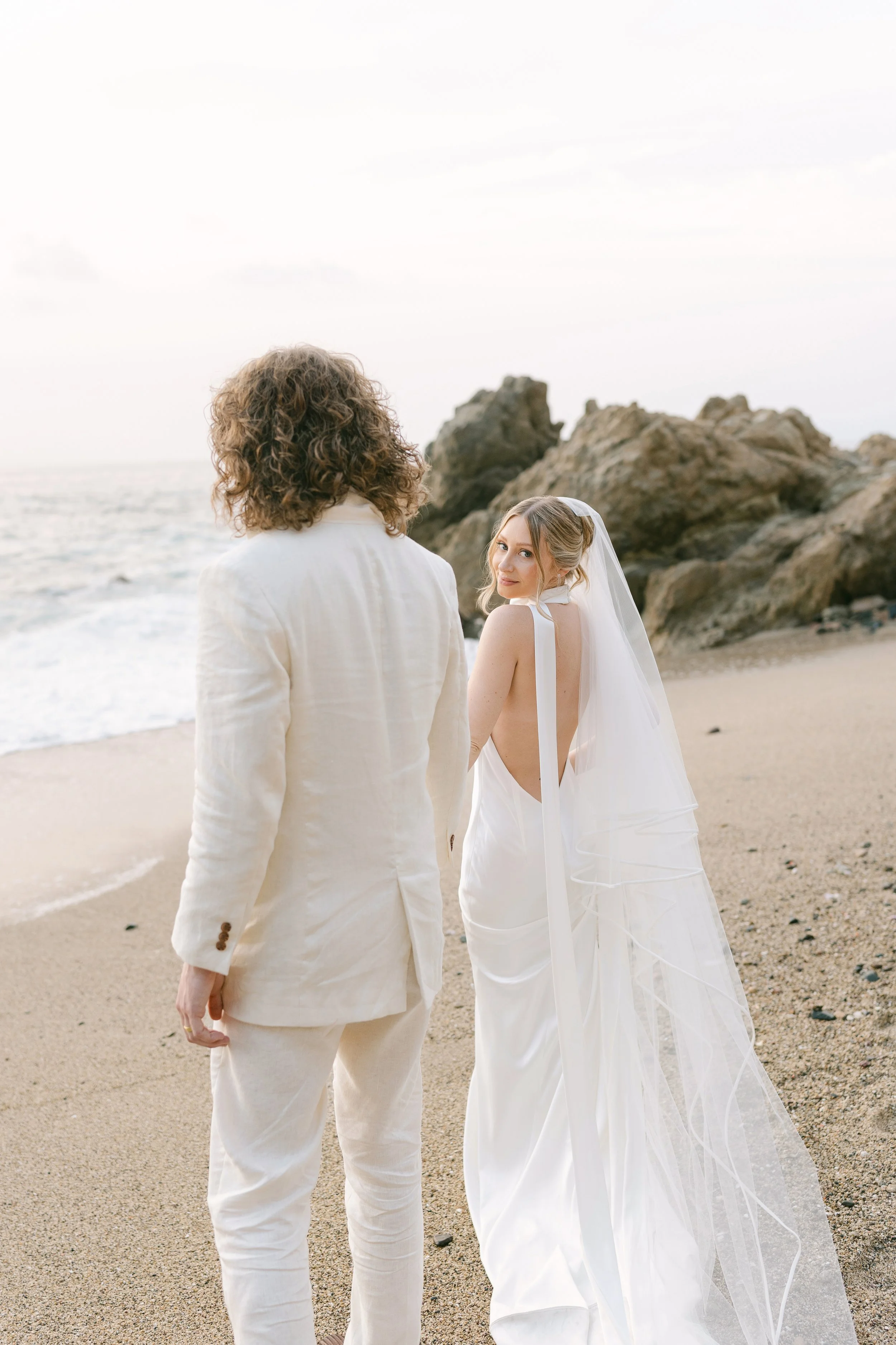 Pareja en ceremonia de boda en una playa con rocas al fondo, vestido de novia y traje de novio en colores claros.