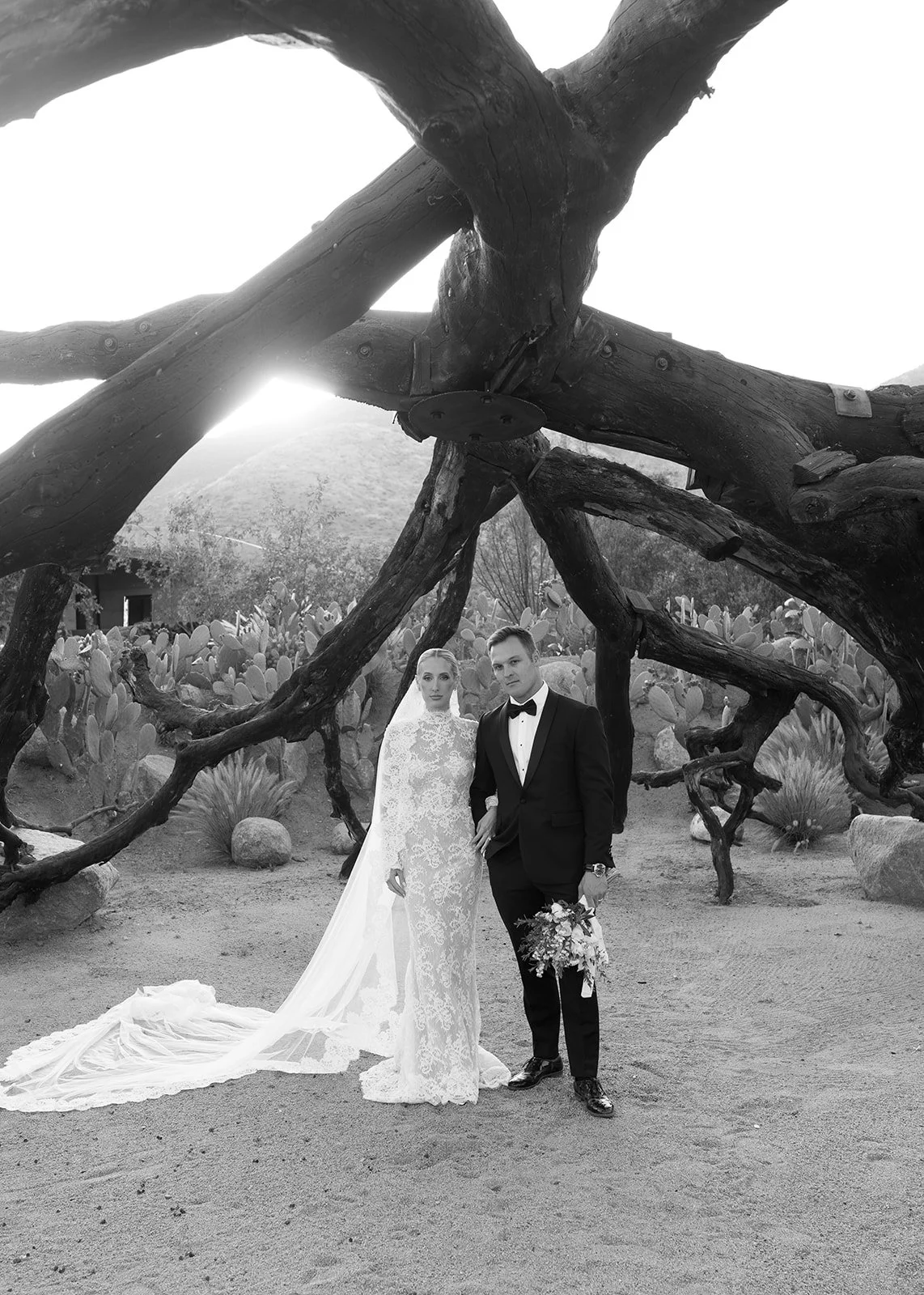 Pareja de novios en traje y vestido de boda, posando en un jardín con cactus y una estructura de madera grande en el fondo, en blanco y negro.