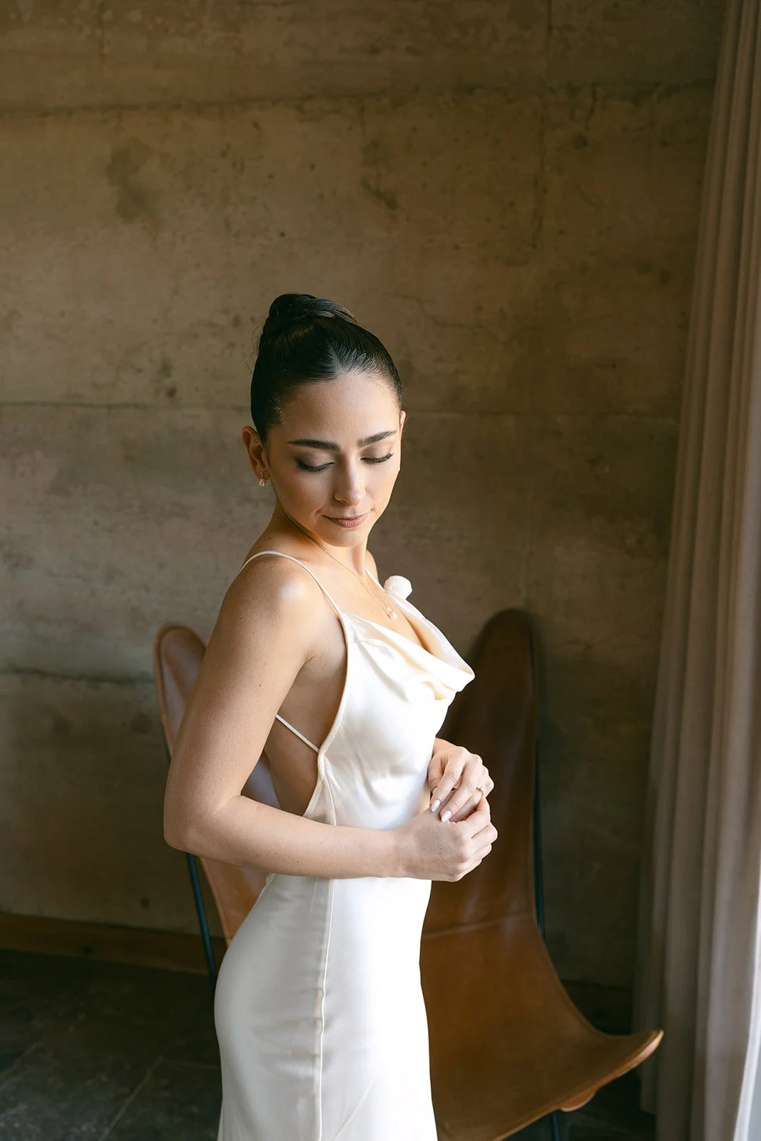 Mujer con vestido de gala blanco, levantando suavemente sus manos en un entorno con pared de piedra y cortinas beige.