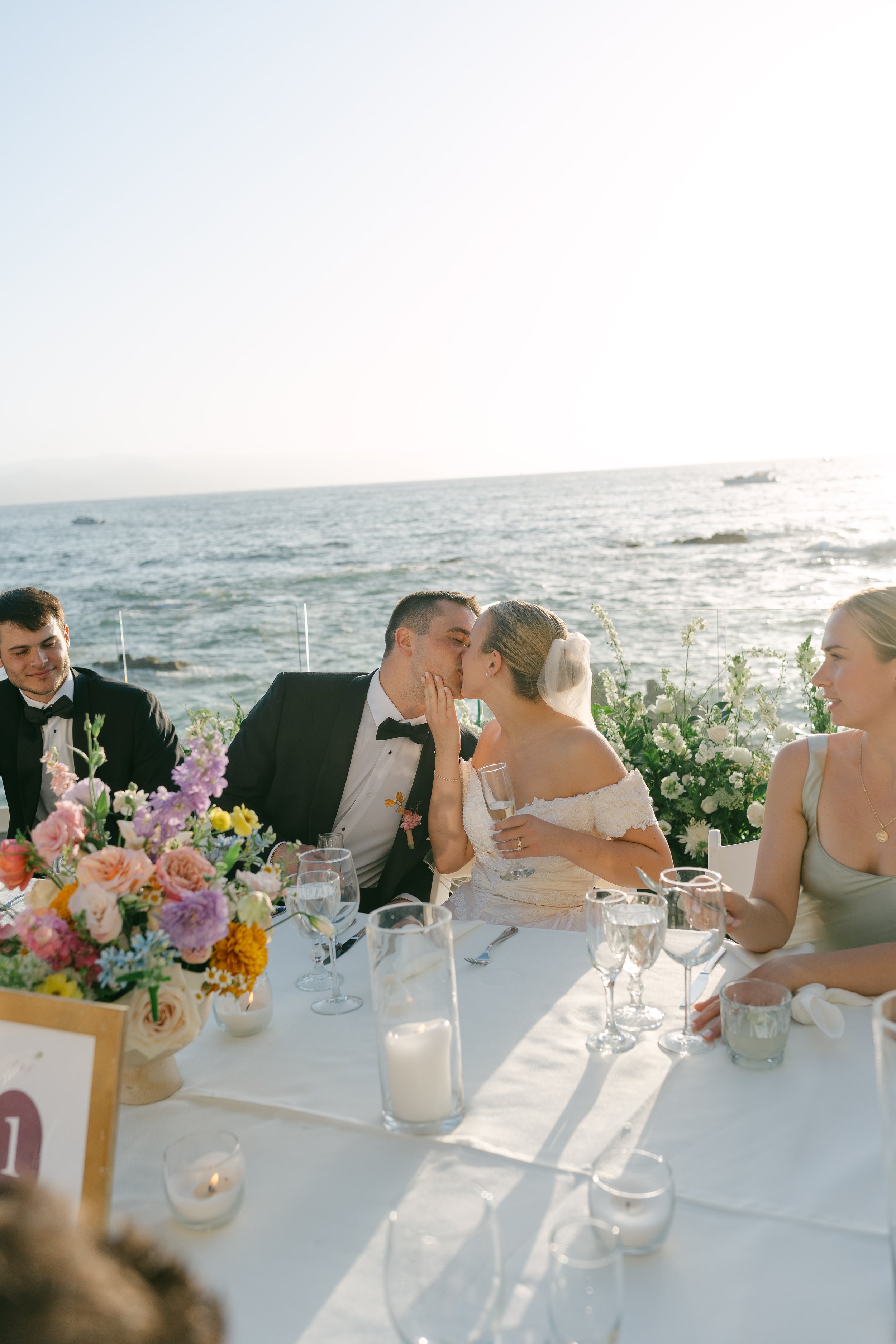 Pareja de novios besándose en una boda junto al mar, rodeados de amigos y flores.