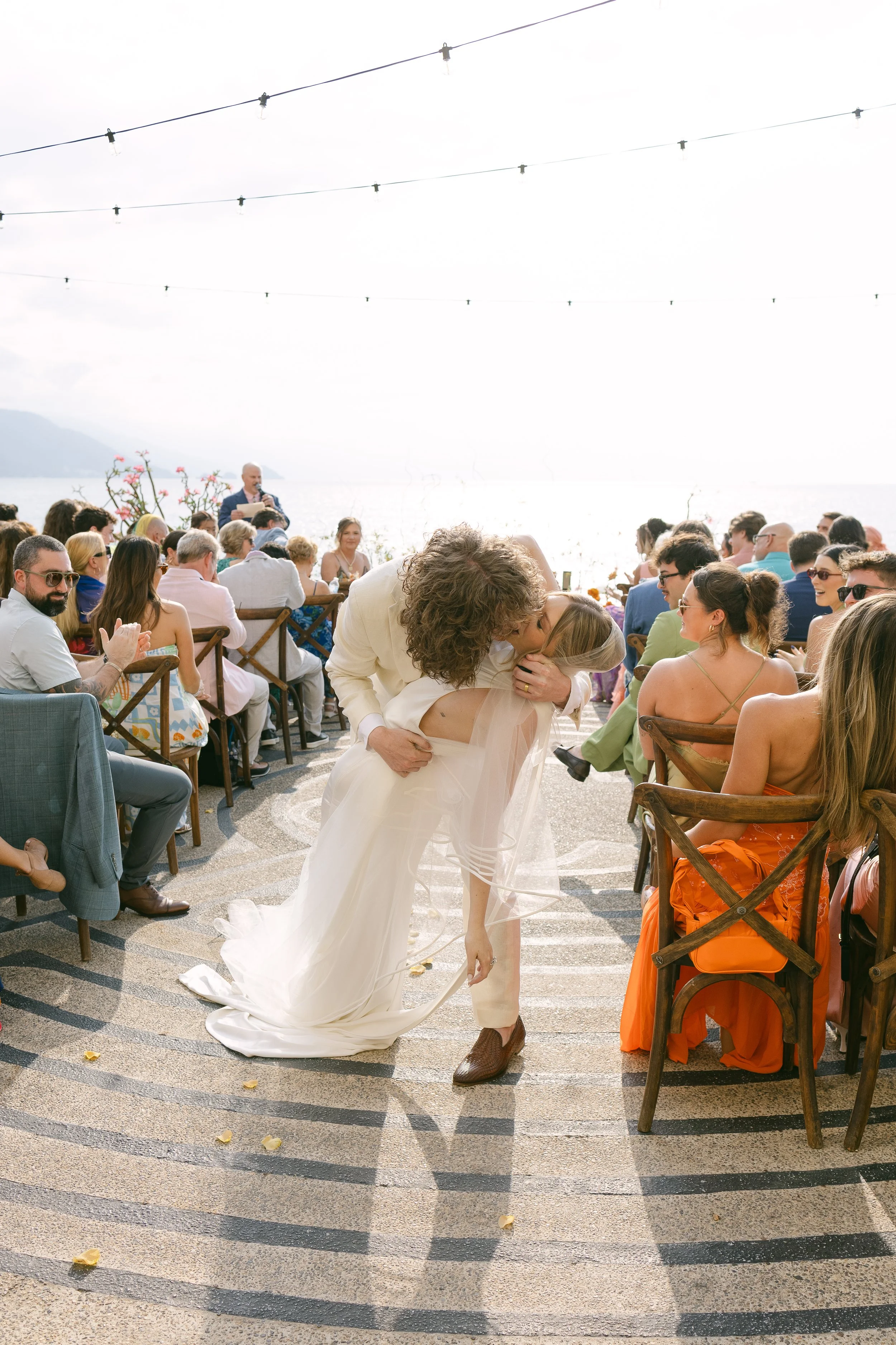 Pareja de recién casados en la playa, bailando y besándose, rodeados de invitados en una ceremonia al aire libre, con el mar y montañas de fondo, en un día soleado.