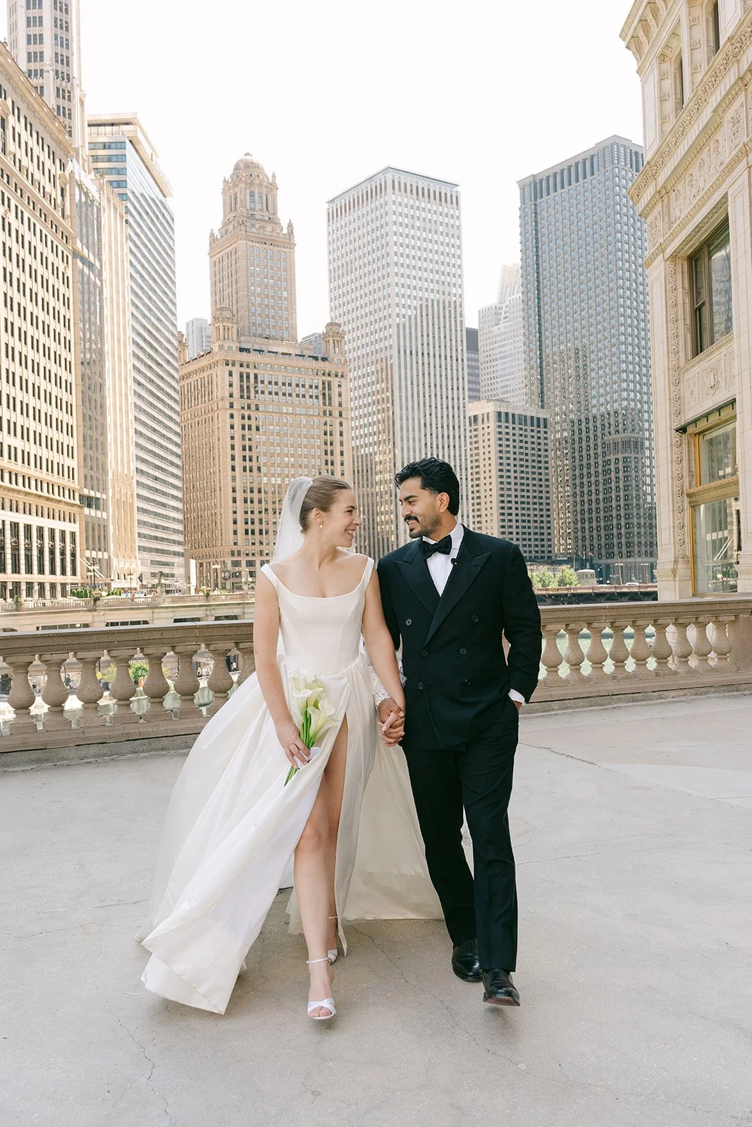 Pareja de recién casados caminando en una terraza con ciudad de rascacielos al fondo, ella con vestido de novia y él con traje y pajarita.
