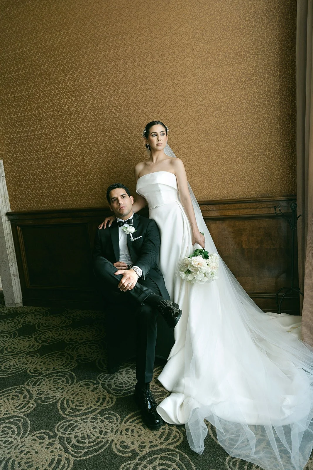 Pareja de recién casados en un ambiente elegante. La novia lleva vestido blanco de sirena con velo y bouquet de flores blancas y crema. El novio viste esmoquin negro con pajarita y flor en el saco. Ella está de pie, apoyando una mano en el hombro del novio, quien está sentado con las piernas cruzadas. Fondo de pared con papel tapiz dorado y decoración clásica.