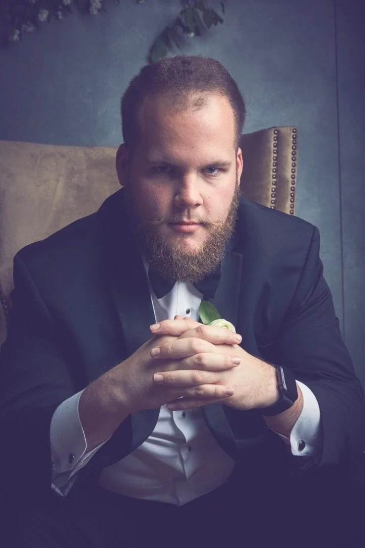 A man in a tuxedo with a beard sitting in a chair with his hands clasped together, looking at the camera.