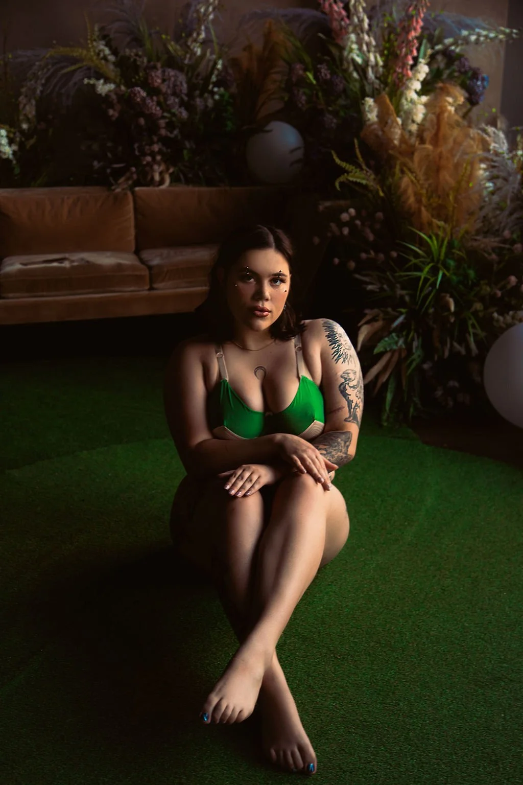 A woman with tattoos wearing a green top, sitting on a green carpet with crossed legs, in front of a backdrop with a brown sofa and large floral arrangements.