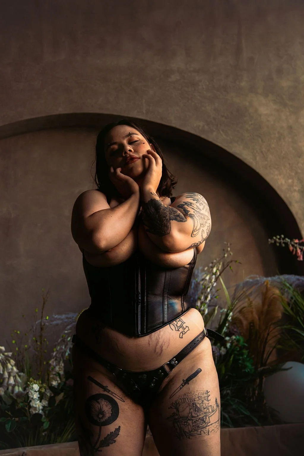 A tattooed woman in black lingerie standing against a neutral background surrounded by flowers, with her eyes closed and one hand touching her face.