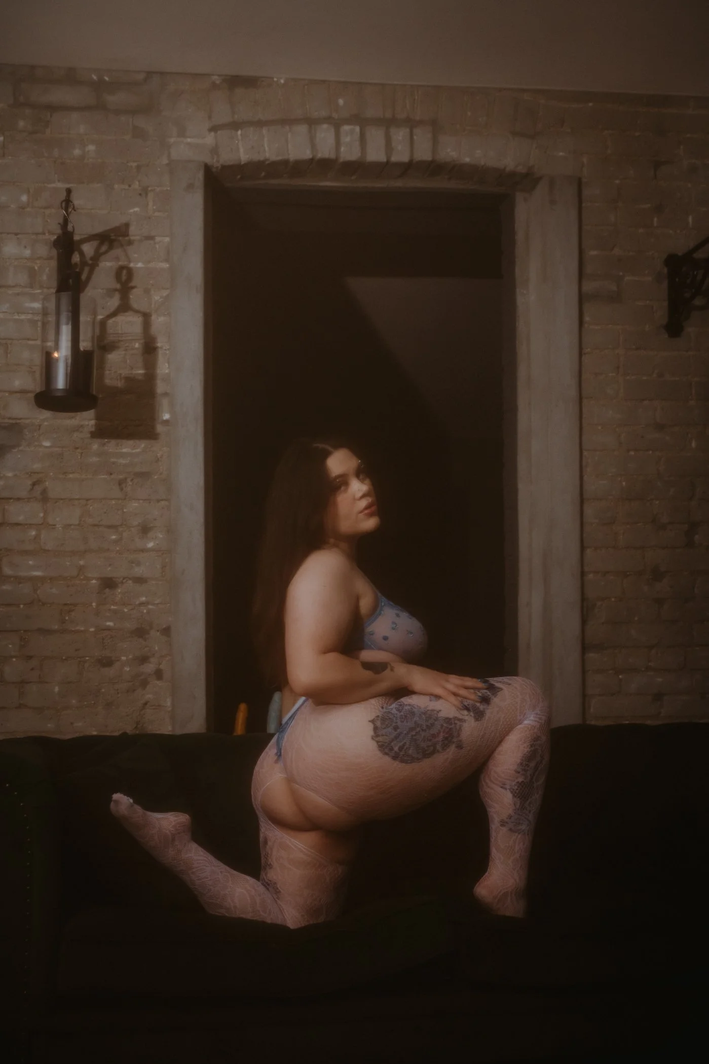 A woman with long dark hair, dressed in sheer stockings and a camisole, kneeling on a black sofa in front of a brick wall with a window opening, with dim lighting.