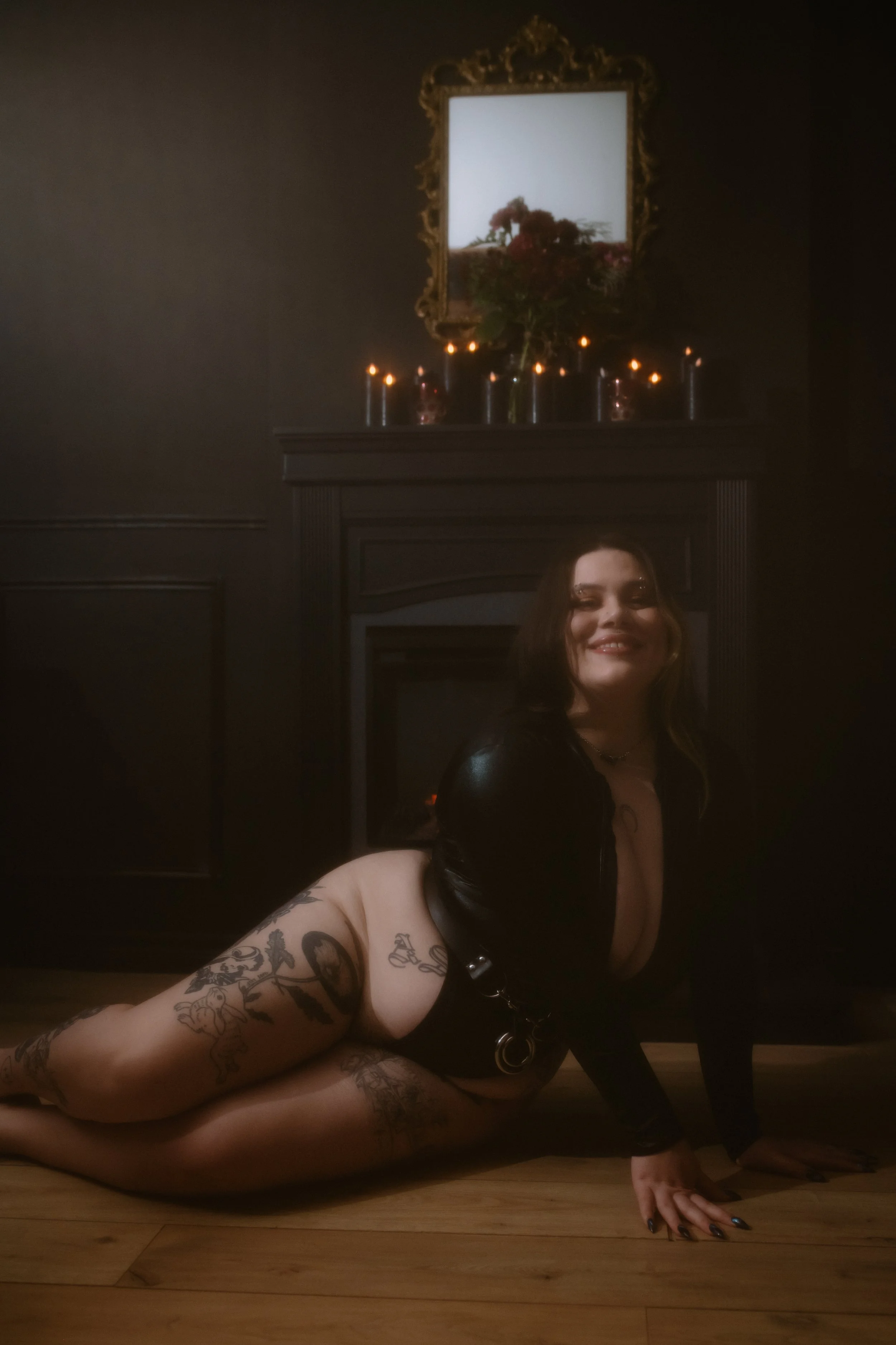 A woman with black hair and tattooed legs poses on a wooden floor in front of a fireplace with a mirror and candles on top, in a dimly lit room.