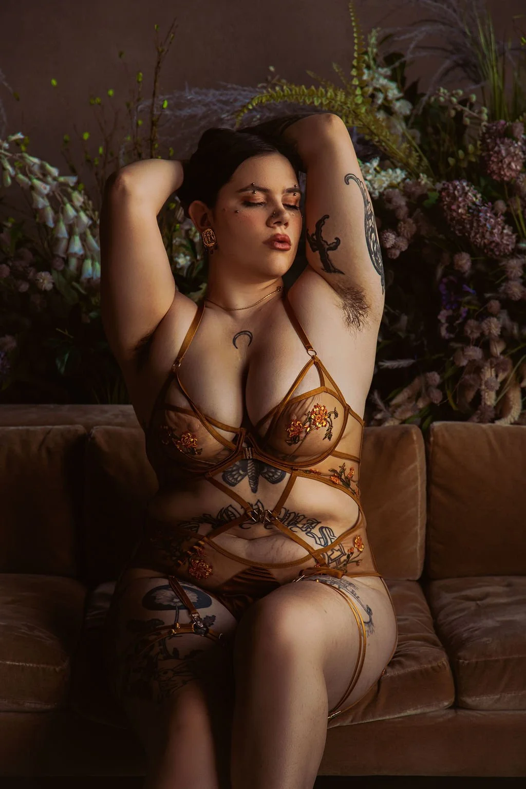A woman with tattoos wearing sheer, embroidered lingerie, sitting on a brown couch with eyes closed and arms raised, surrounded by flowers and greenery.
