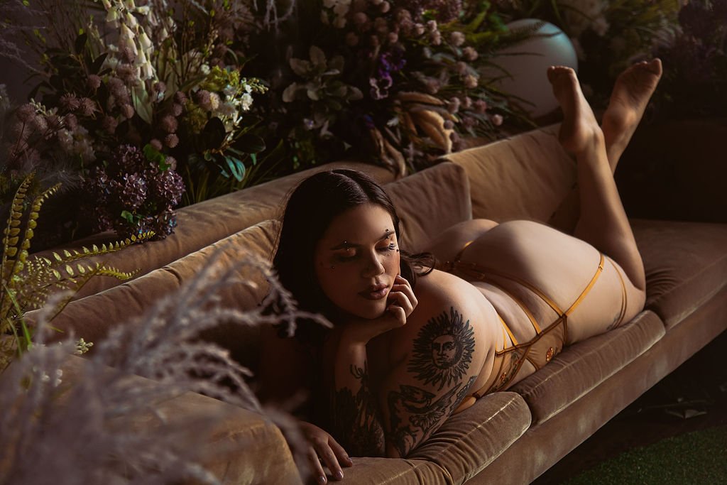 A woman with dark hair and tattoos lying on a beige velvet couch surrounded by flowers and greenery, resting her head on her hand with her legs bent and raised.