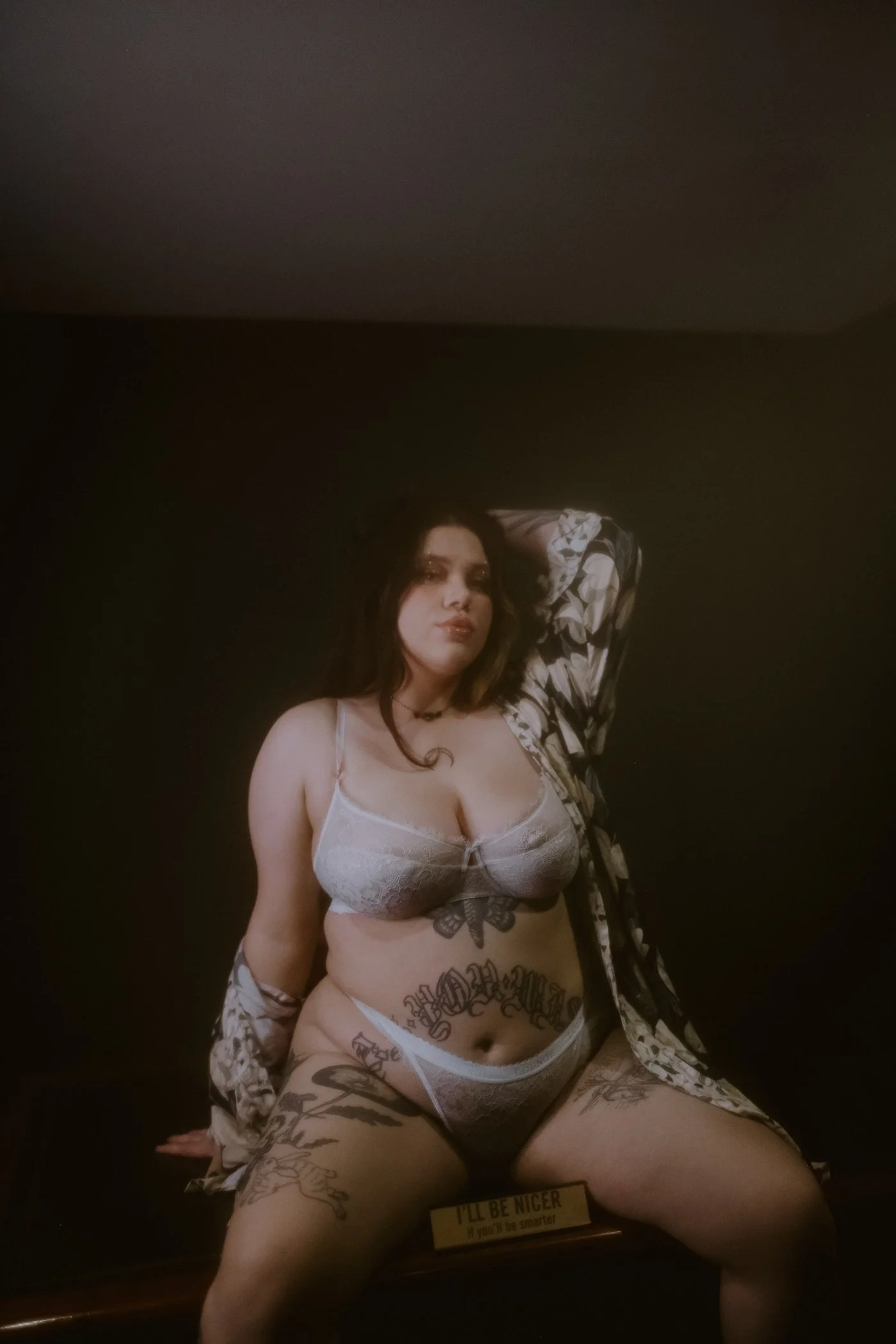 A woman wearing white lingerie, sitting against a dark background, with tattoos on her arms, stomach, and thighs, and a black choker around her neck.