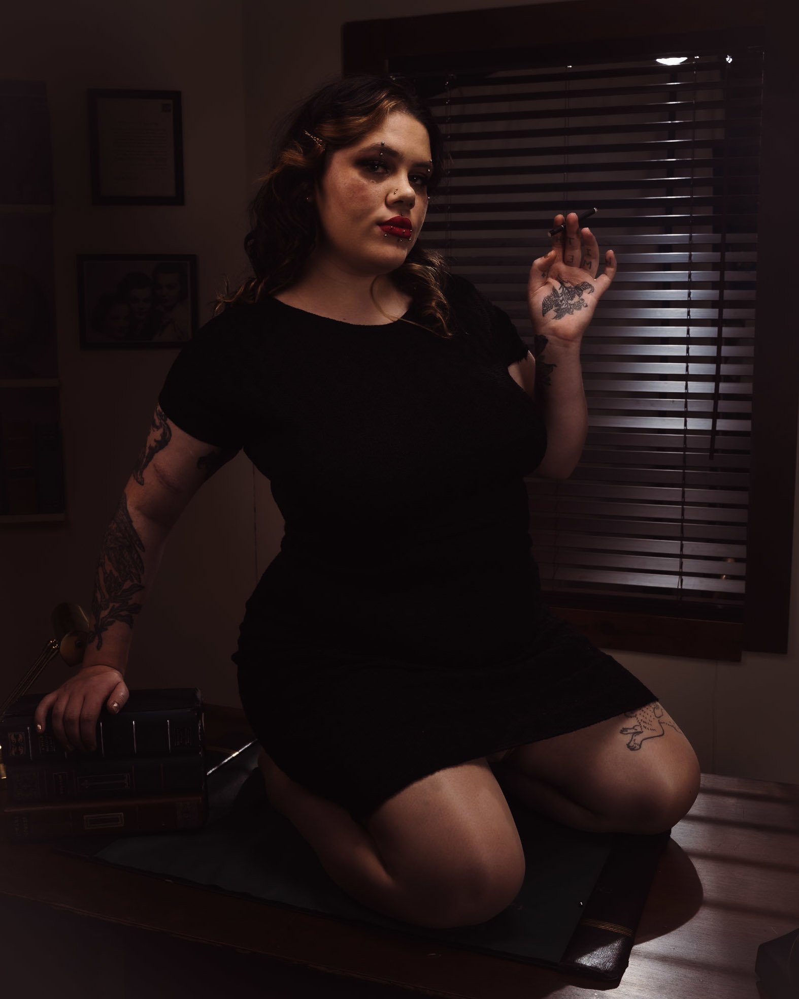 A woman with tattoos and dark hair holding a cigarette, wearing a black dress, kneeling on a table in a dimly lit room.