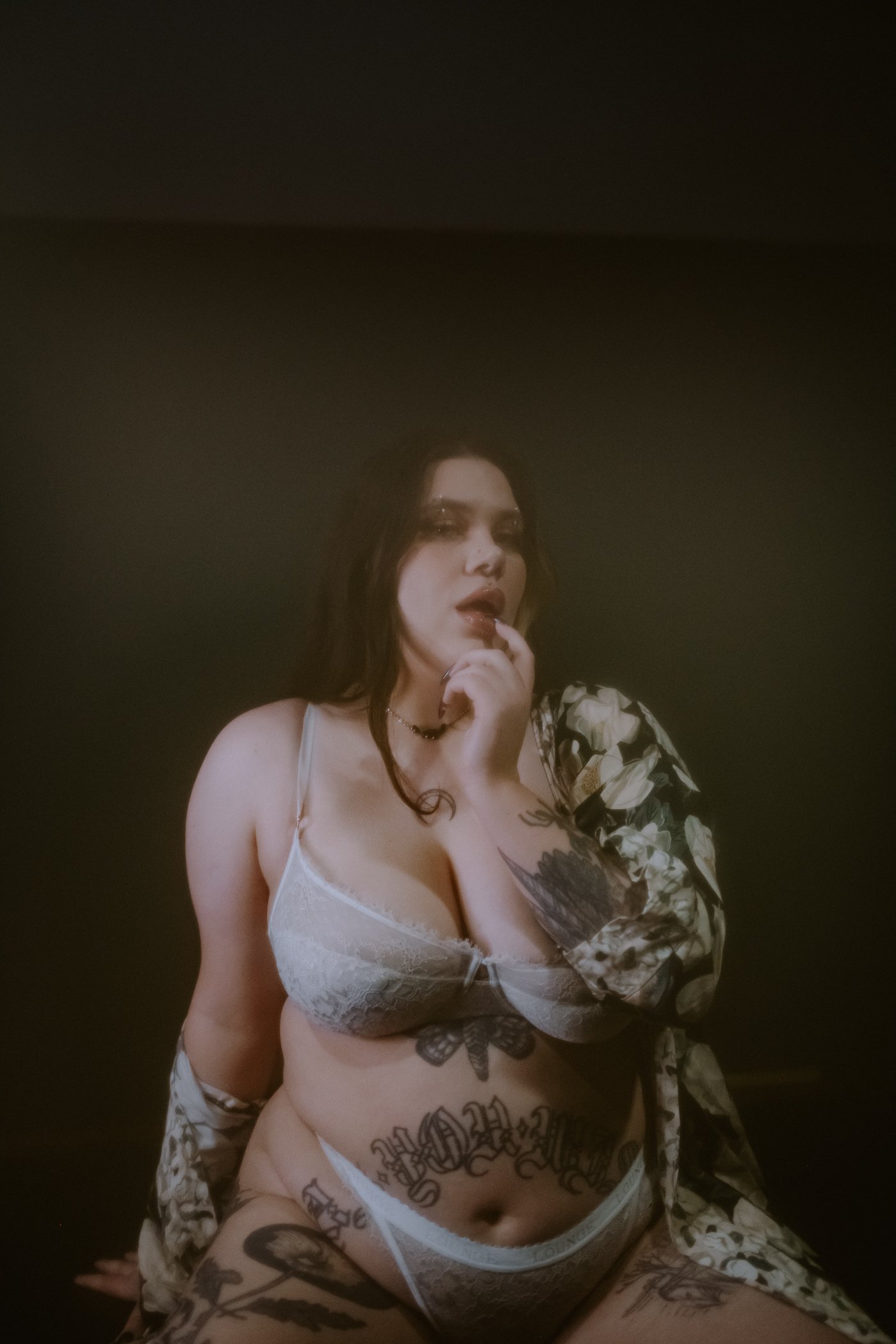 A woman with dark hair, tattoos, wearing white lace lingerie, posing against a dark background.