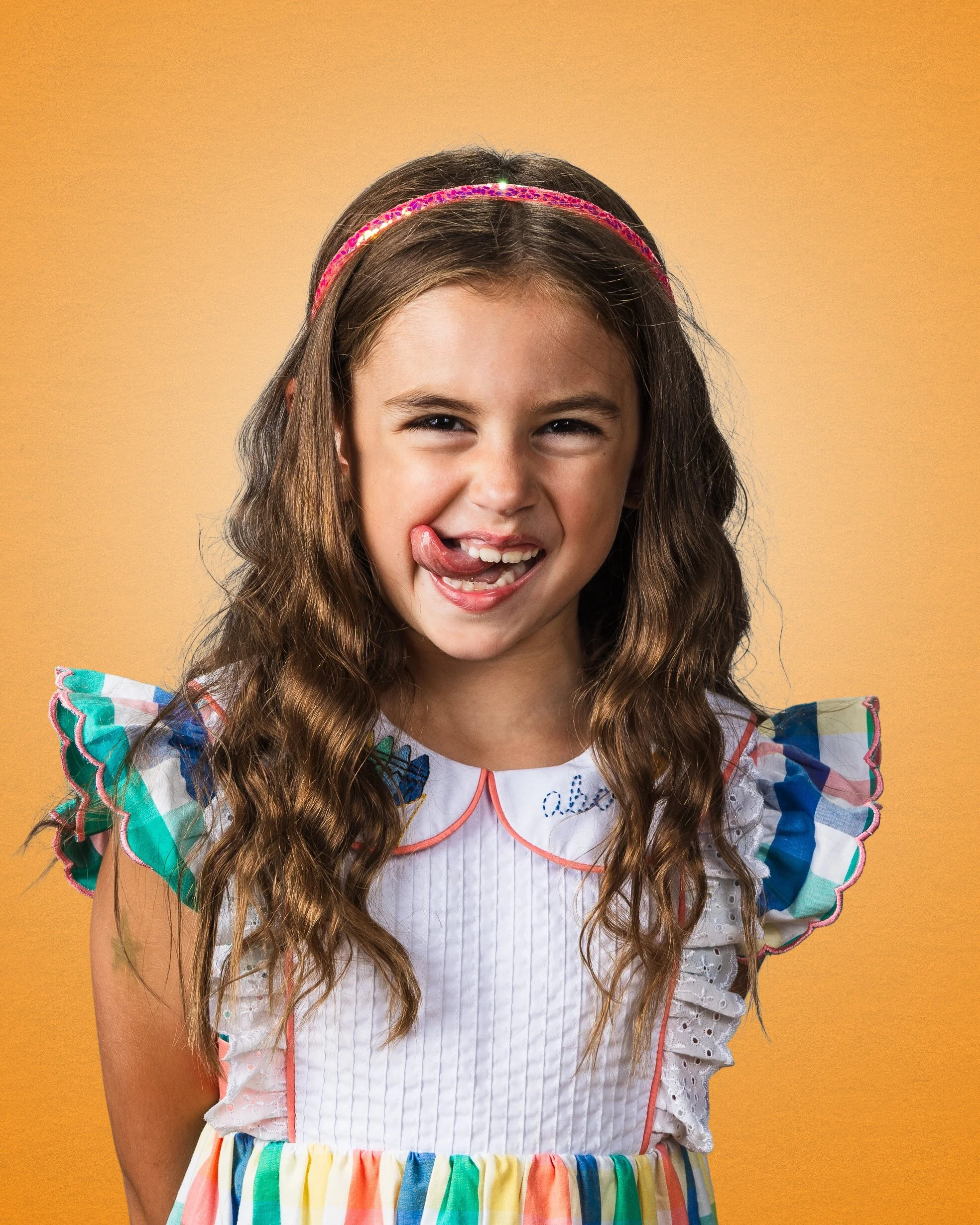 A young girl with long, wavy brown hair and light skin, wearing a colorful dress with ruffled sleeves and a pink headband, sticking out her tongue and smiling, against a yellow background.