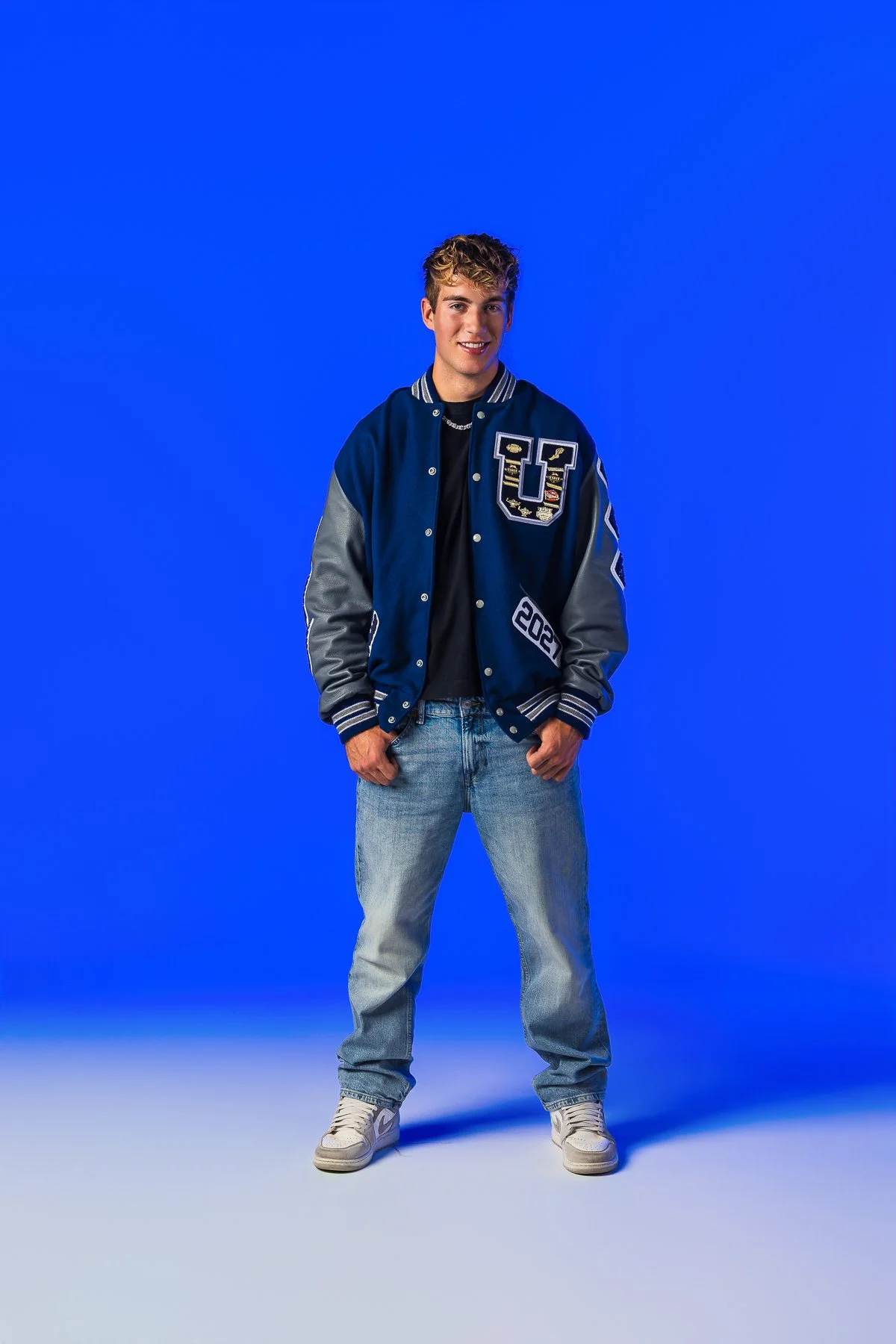 A young man stands smiling against a solid blue background wearing a navy blue varsity jacket with gray sleeves, light blue jeans, and white sneakers.